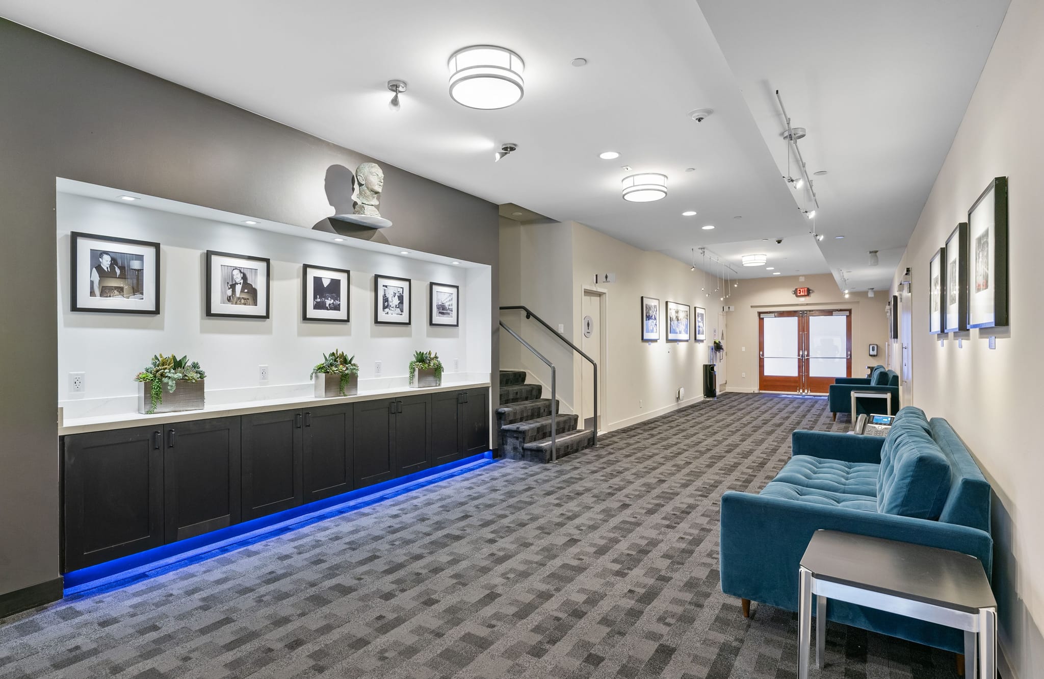 A long, well-lit hallway with a display wall featuring framed photos, a bust, and blue-lit cabinets. Teal sofas line a geometric patterned carpet, with stairs and double doors visible further down.