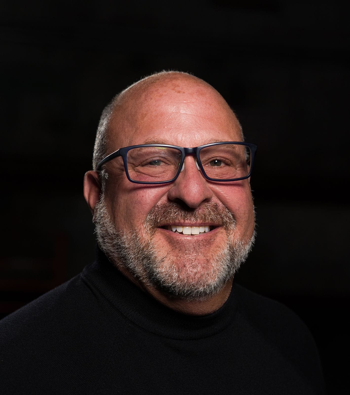 A smiling, bald man with a beard and glasses wears a black turtleneck against a dark background.