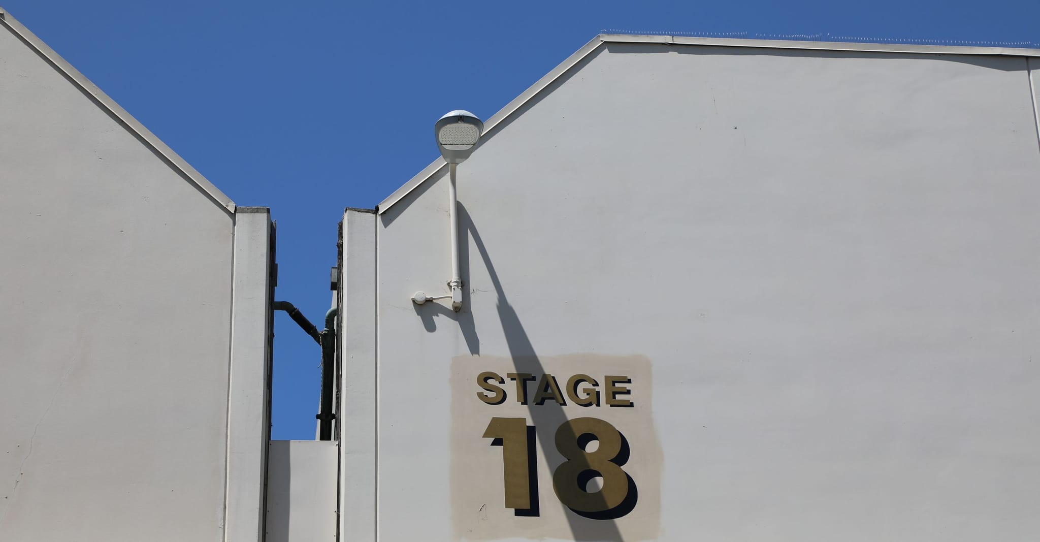 White building wall with "STAGE 18" written on it in large golden letters, under a bright blue sky.