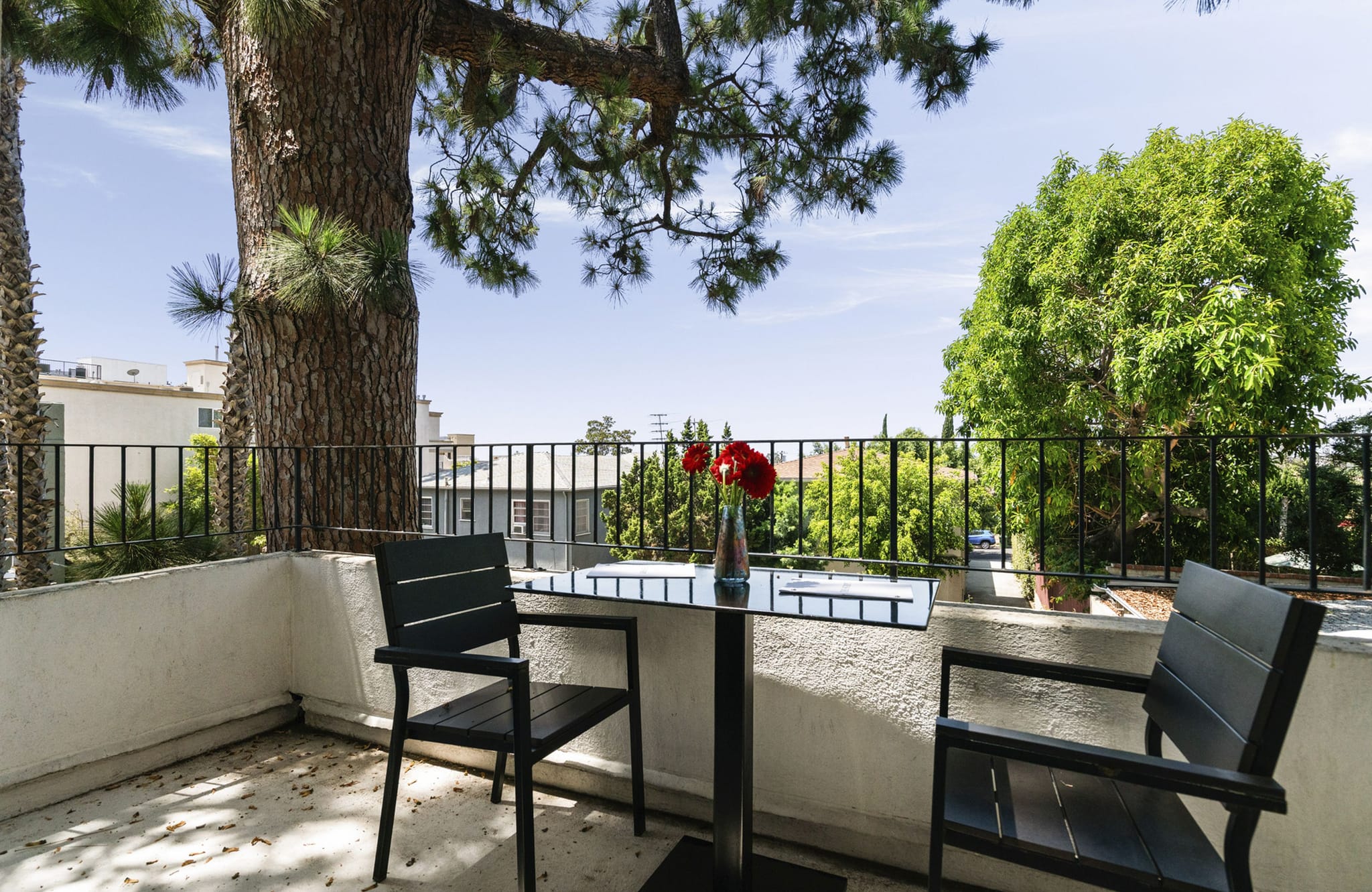A balcony with a small table, two chairs, and a vase of red flowers, surrounded by trees and a railing.