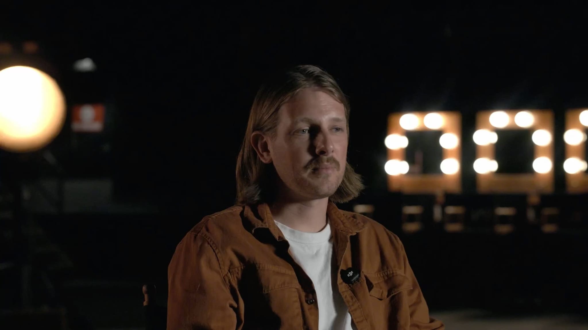 A man with long hair and a mustache wearing a brown shirt in a dimly lit studio setting.