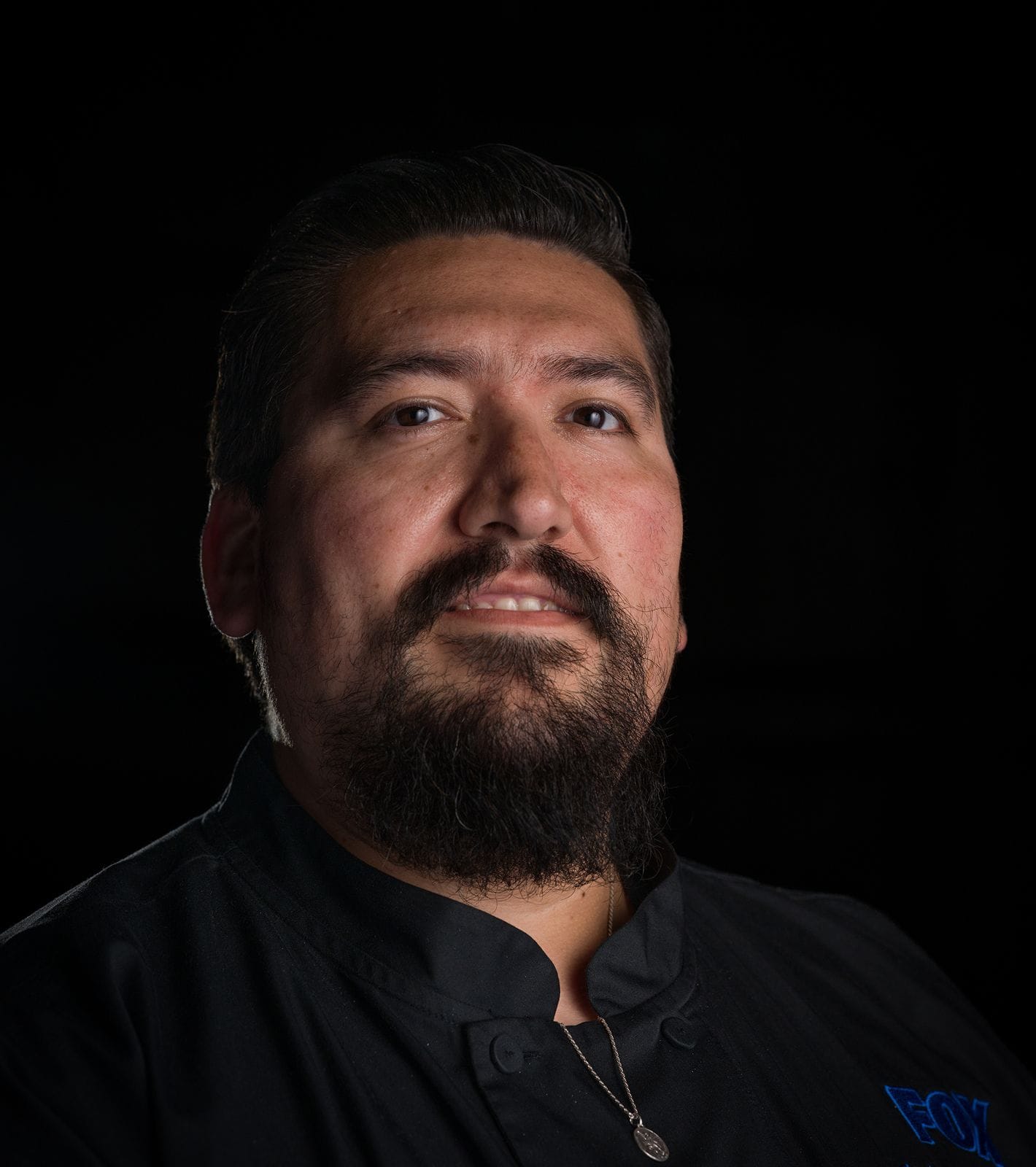 Portrait of a man with dark hair and a beard, wearing a black chef's jacket with a blue FOX logo, smiling against a black background.