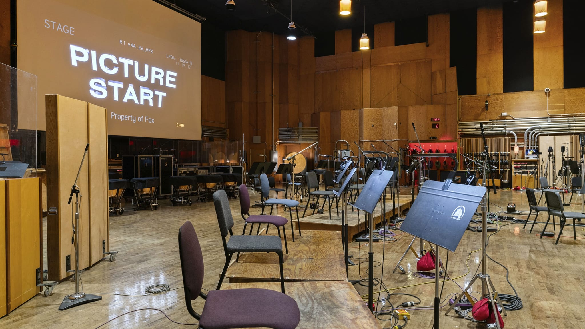 An empty orchestra recording studio with "PICTURE START" projected on a screen, surrounded by chairs, music stands, and microphones.