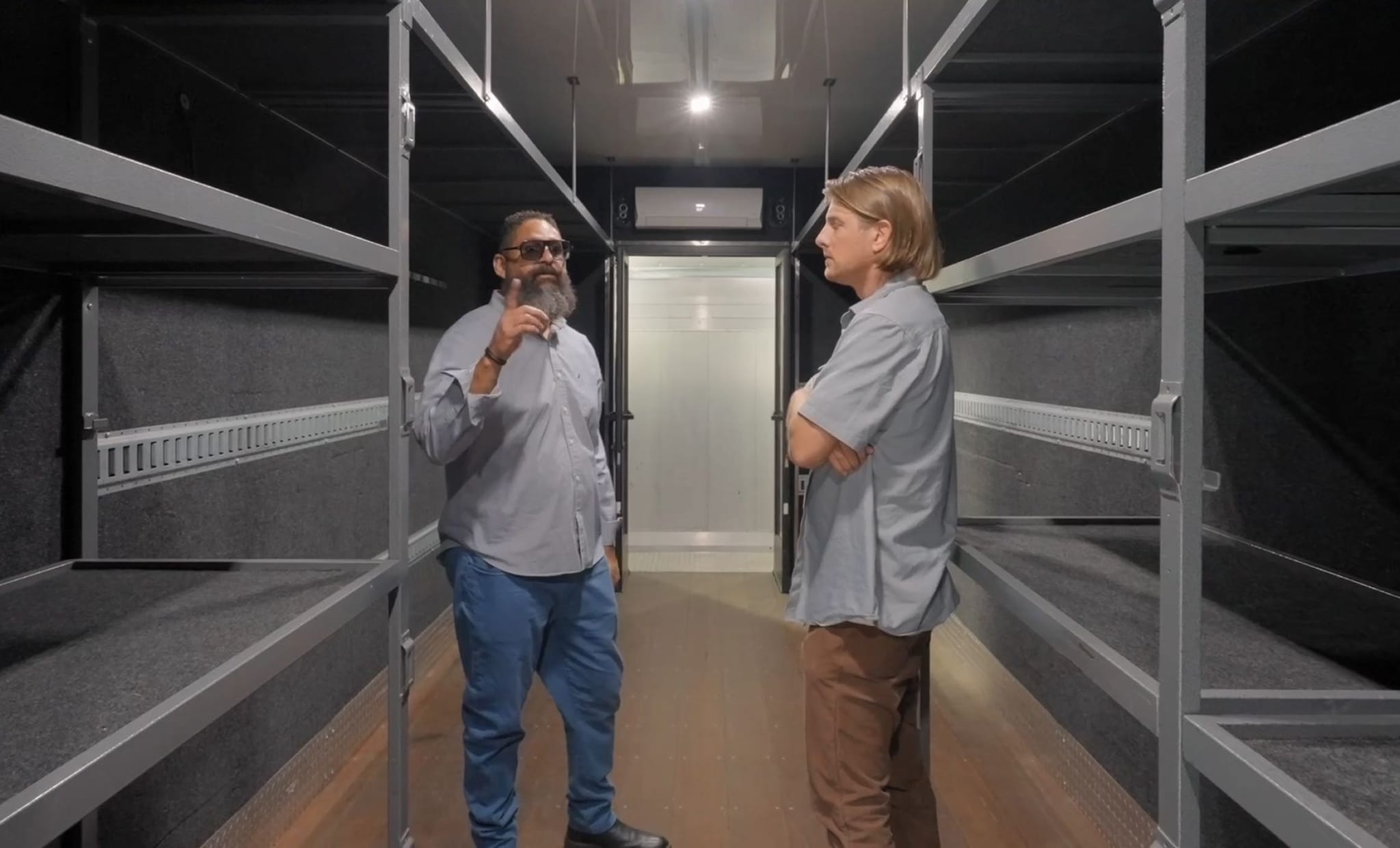 A bearded man in sunglasses gestures to a blonde man with crossed arms, inside a dark, multi-level bunk or storage unit.