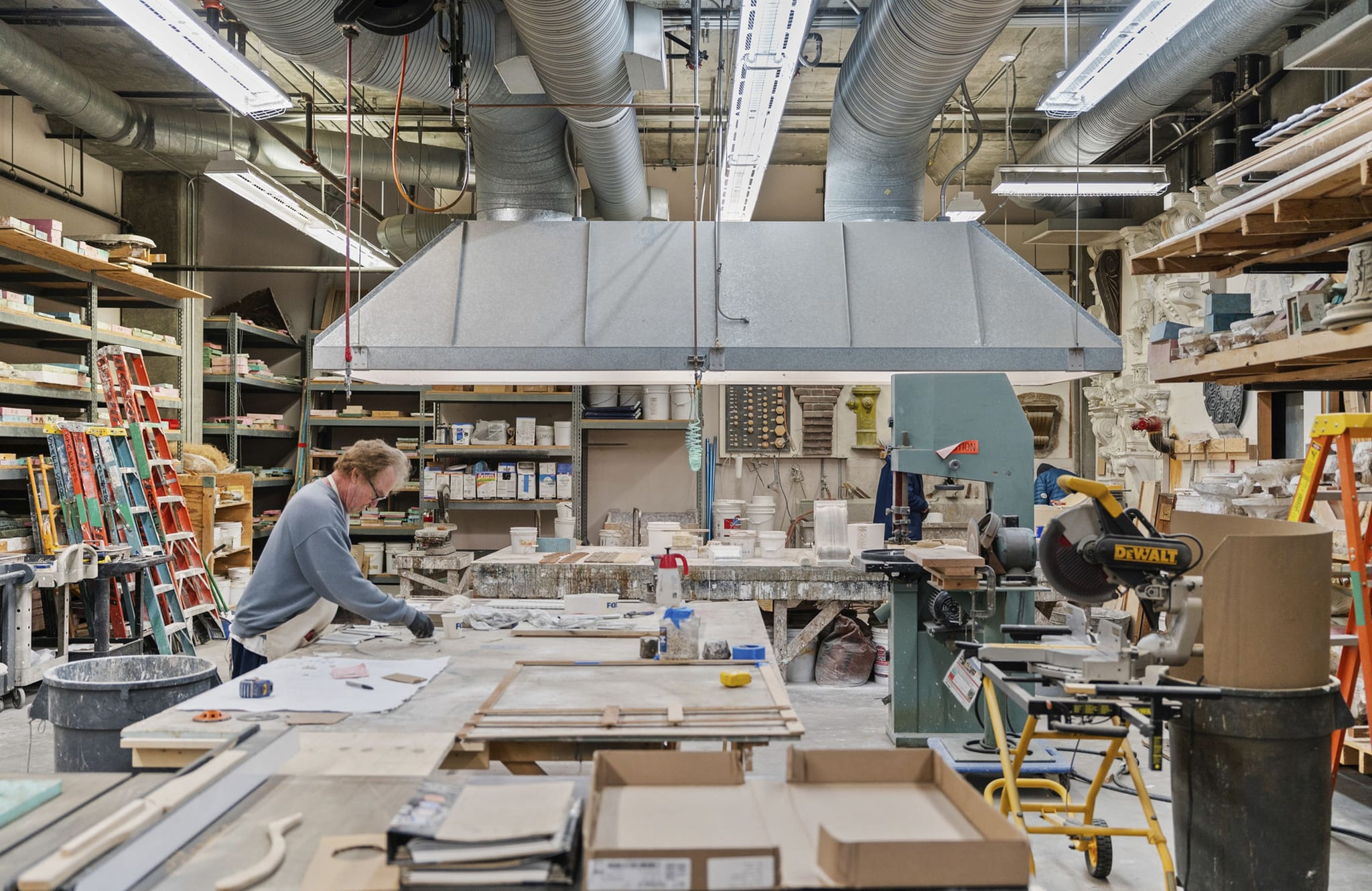 A man works at a workbench in a large industrial workshop filled with tools and materials.