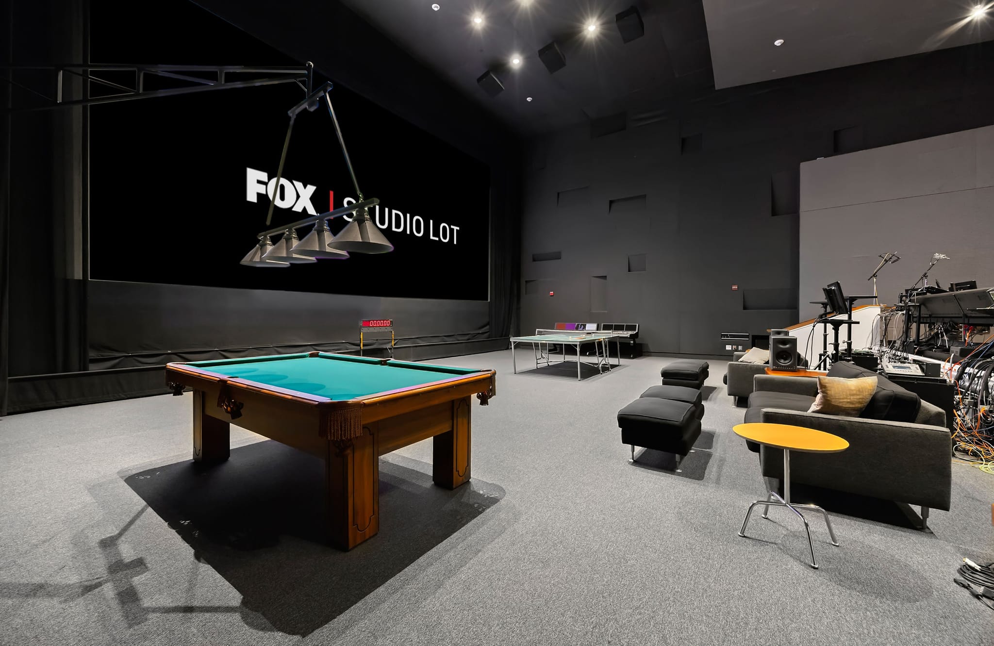 A large, dark recreational room with a screen showing "FOX STUDIO LOT", a pool table, and lounge furniture.