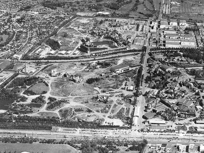 Aerial black and white photo of Disneyland under construction, with Sleeping Beauty Castle, open ground, roads, and parking lots.