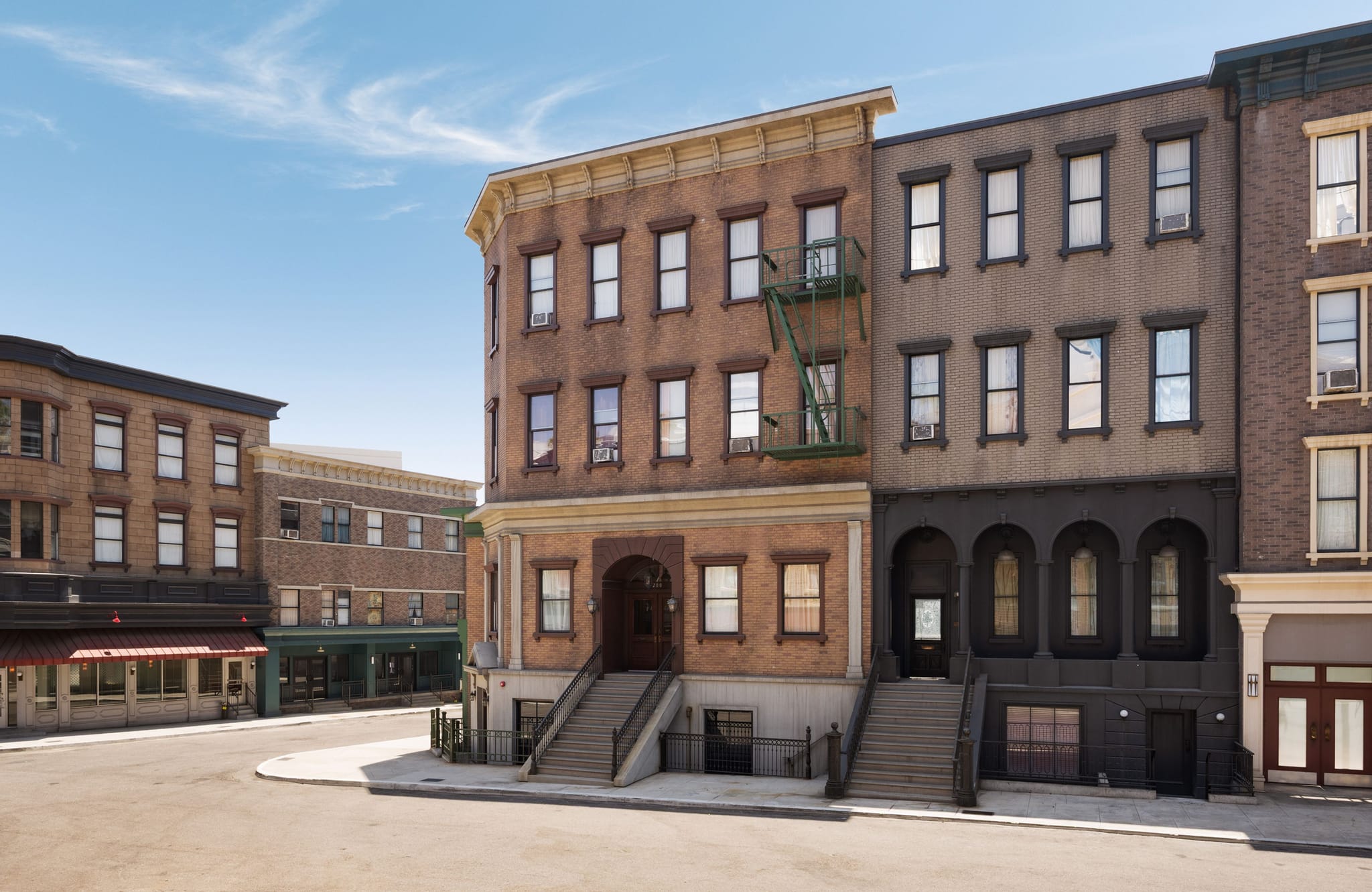 An empty street corner lined with multi-story urban buildings, one with a fire escape, under a blue sky.