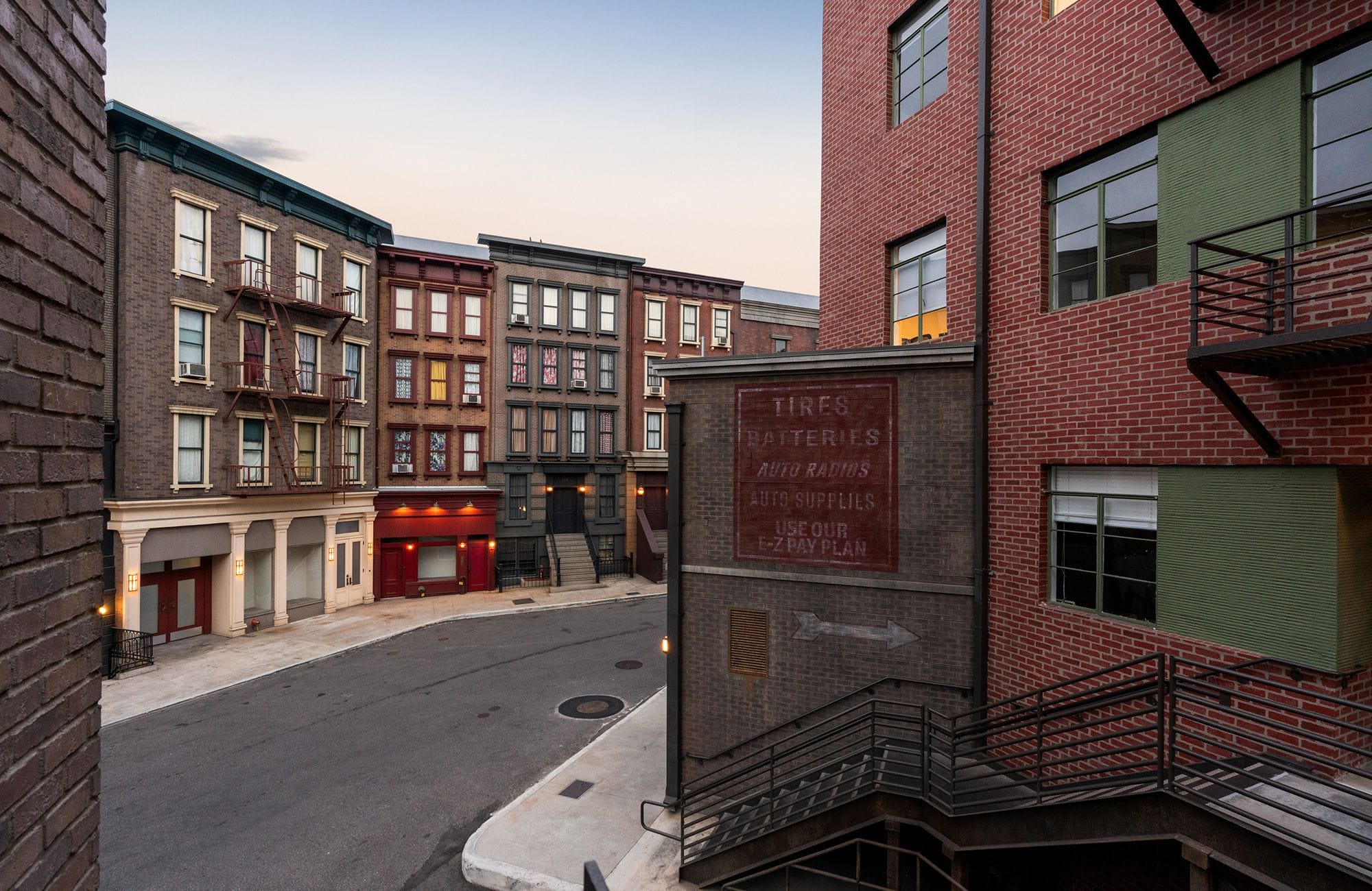 A quiet street lined with old brick buildings, some with fire escapes, and a large weathered "TIRES BATTERIES" sign on a corner building.