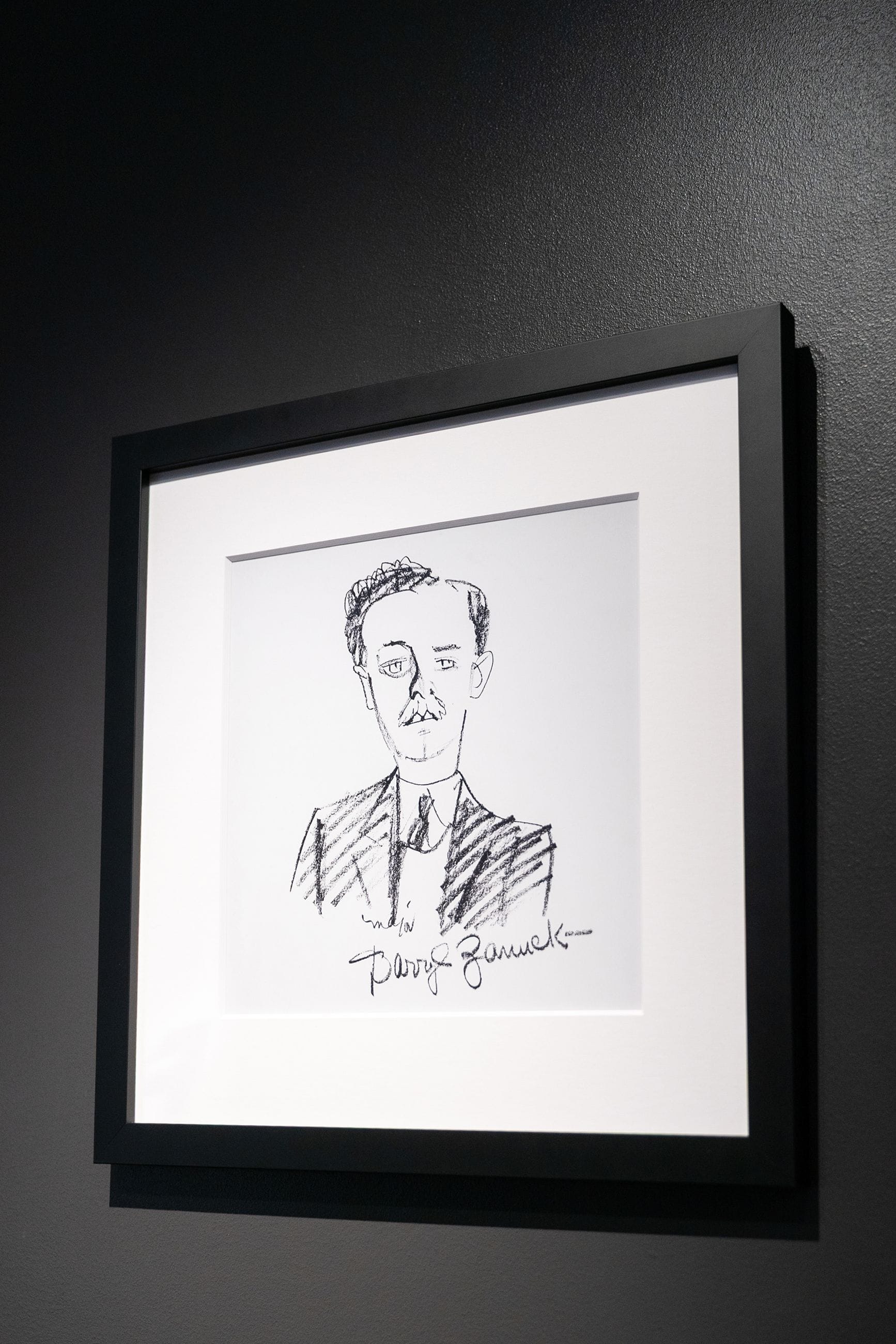 Framed black and white caricature of a man with a mustache and suit, signed 'Darryl Zanuck', on a dark wall.