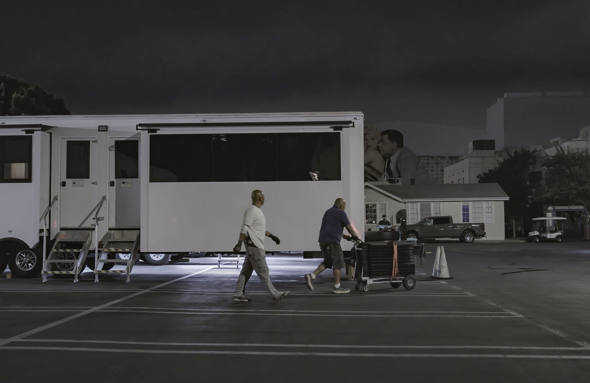 Night scene in a parking lot with a large white production trailer, two men walking, and a grayscale image of a kissing couple in the background.