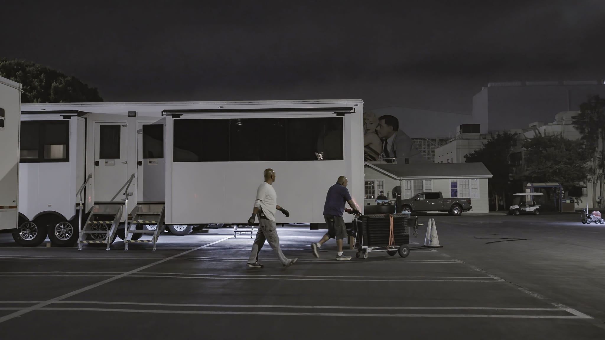 Two men walk past a large white production trailer in a dimly lit parking lot at night, with a blurry image of a man's face visible in the background.