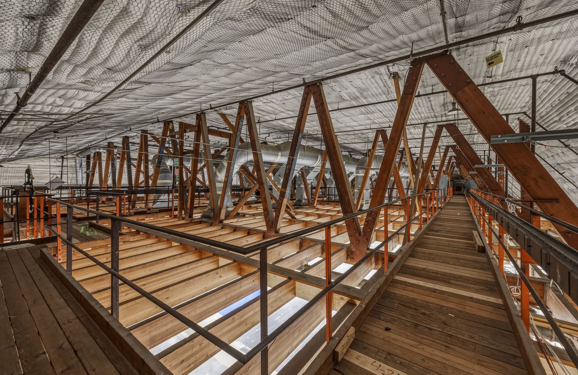 Industrial interior with a dense network of wooden A-frame trusses, exposed ducts, a wooden walkway, and an insulated ceiling.