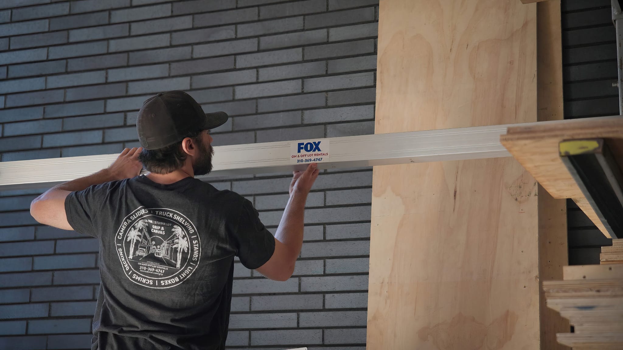 A man holds a silver beam with a "FOX" logo against a gray brick wall.