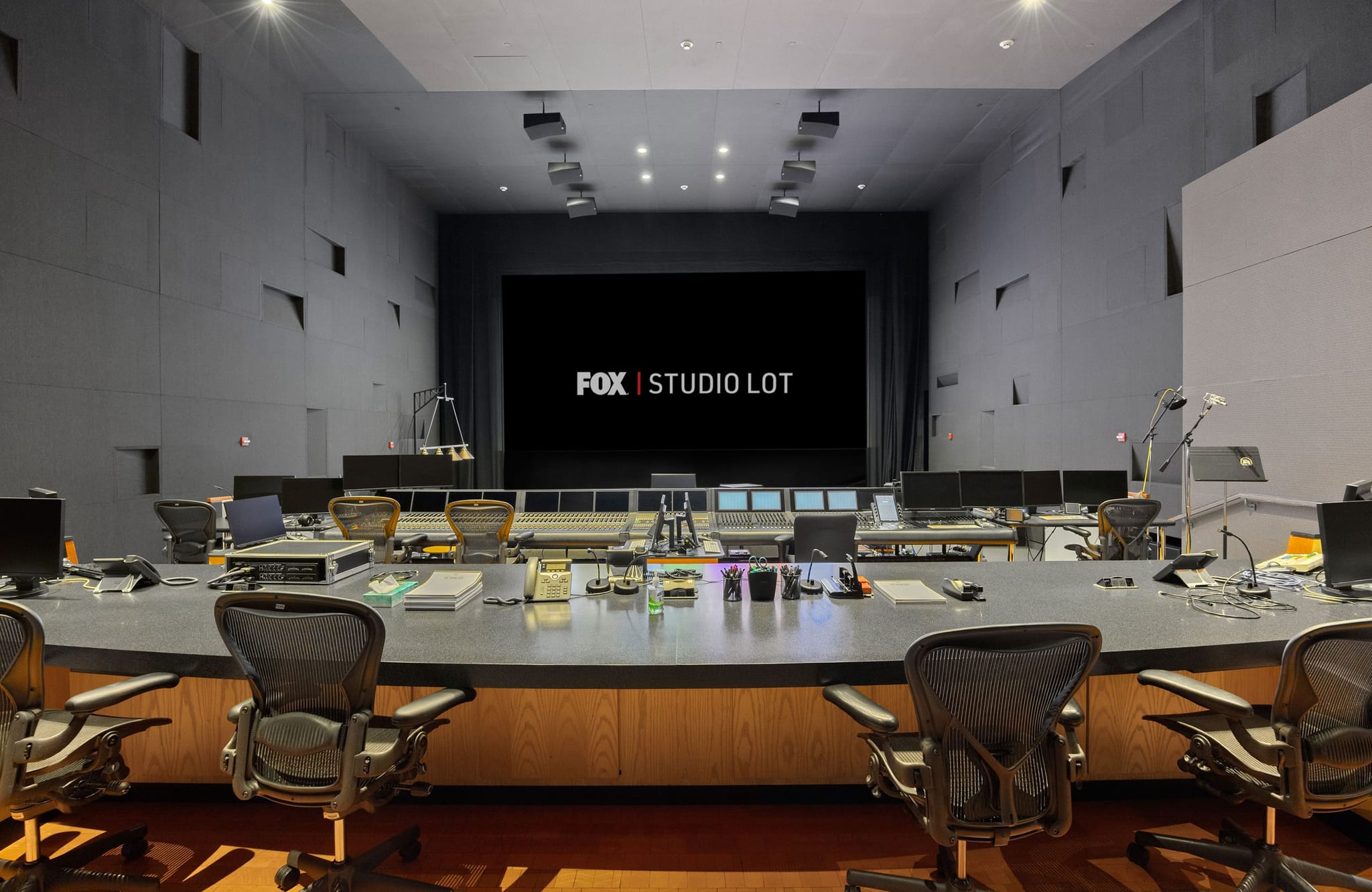 A modern media control room with a curved console and chairs, facing a large black screen displaying "FOX | STUDIO LOT."