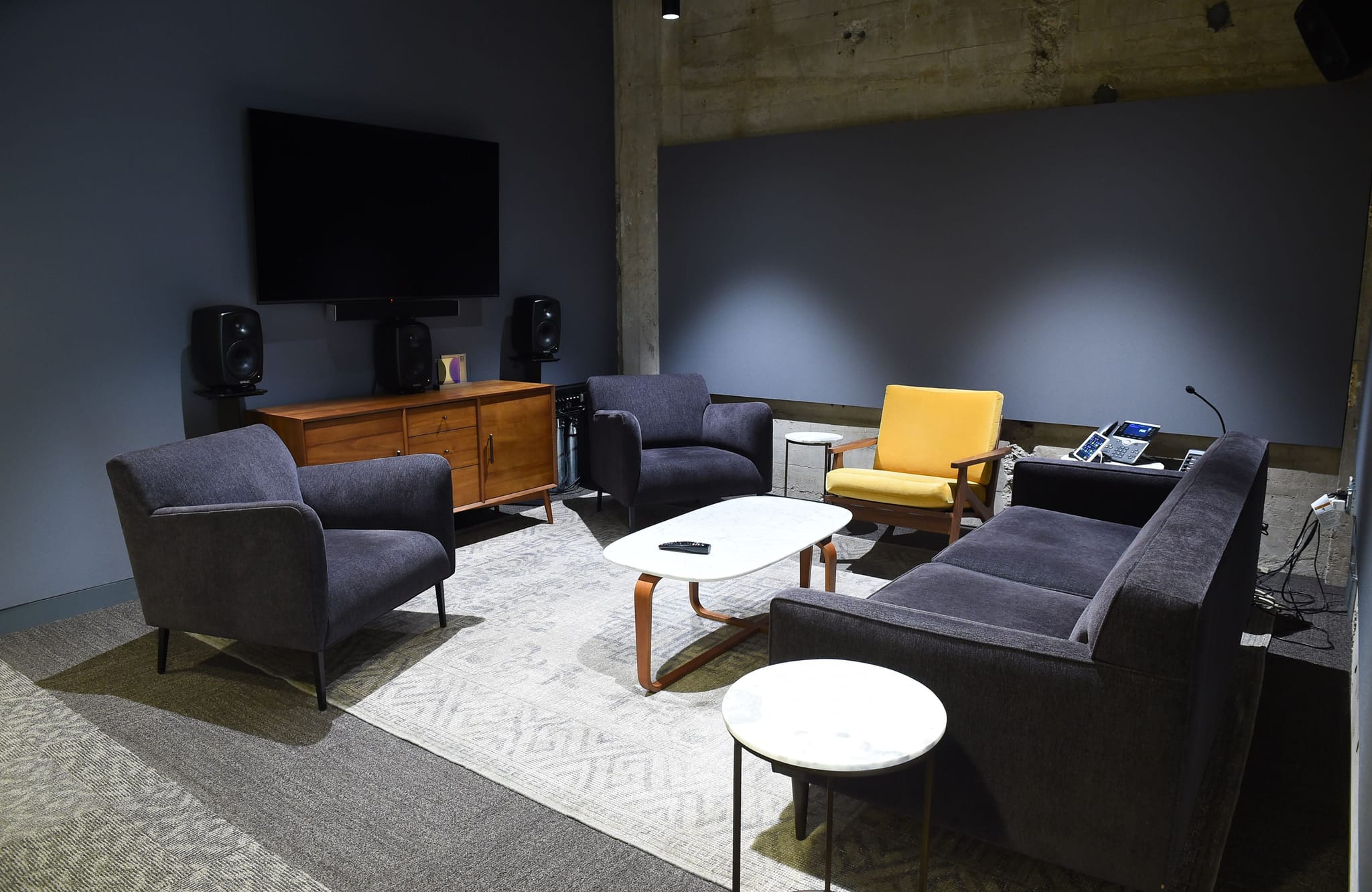 A modern lounge with a TV, wooden credenza, two gray armchairs, a yellow armchair, a gray sofa, and coffee tables on a patterned rug.
