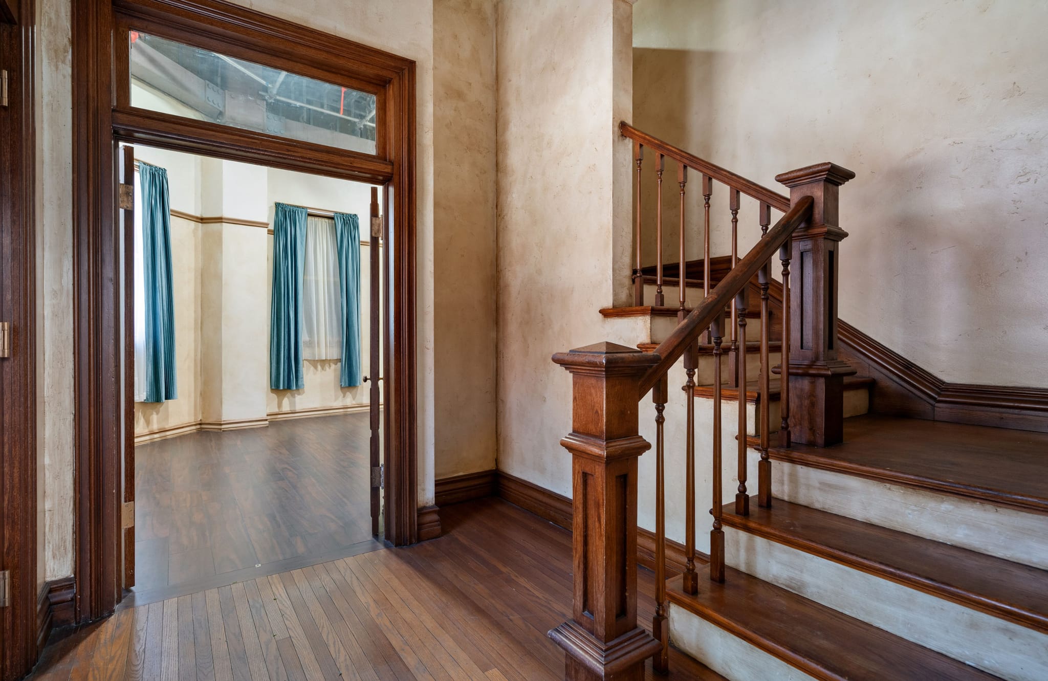 Empty room with dark wood floors, a wooden staircase on the right, and an open doorway to another room with blue curtains.