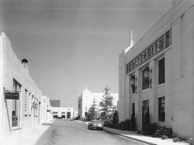 Black and white photo of a street lined with studio buildings, one labeled 'STAGES 1 & 2' and another 'ANIMATION BLDG No 1', with a vintage car.