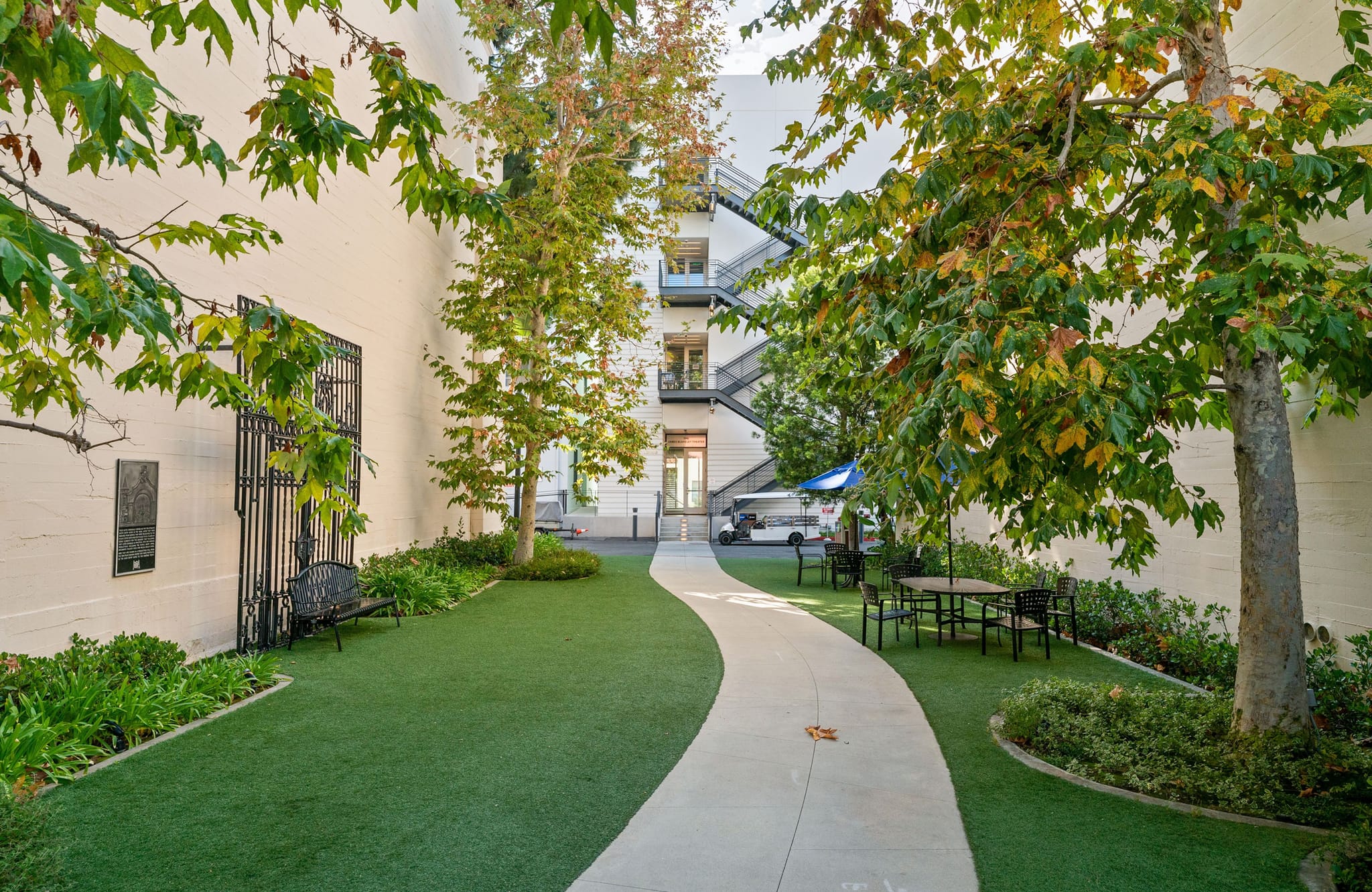A winding concrete path through a green courtyard with trees, a bench, and an outdoor dining set, enclosed by white building walls.