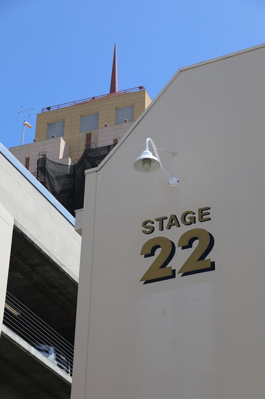 A building wall with "STAGE 22" in gold letters, a white light fixture, and other buildings against a clear blue sky.