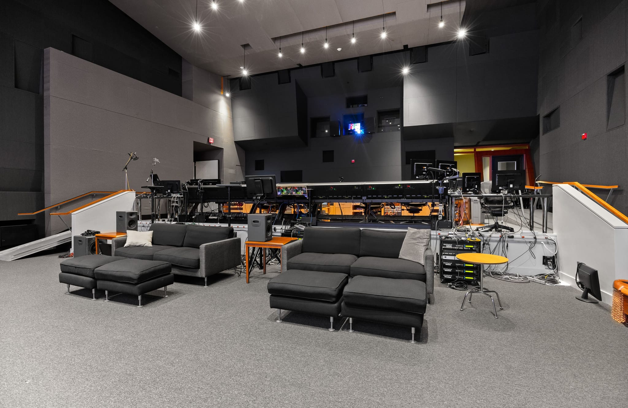 A spacious, modern recording studio with dark gray sound-dampening walls, two sectional sofas, and a long console filled with audio equipment and monitors.