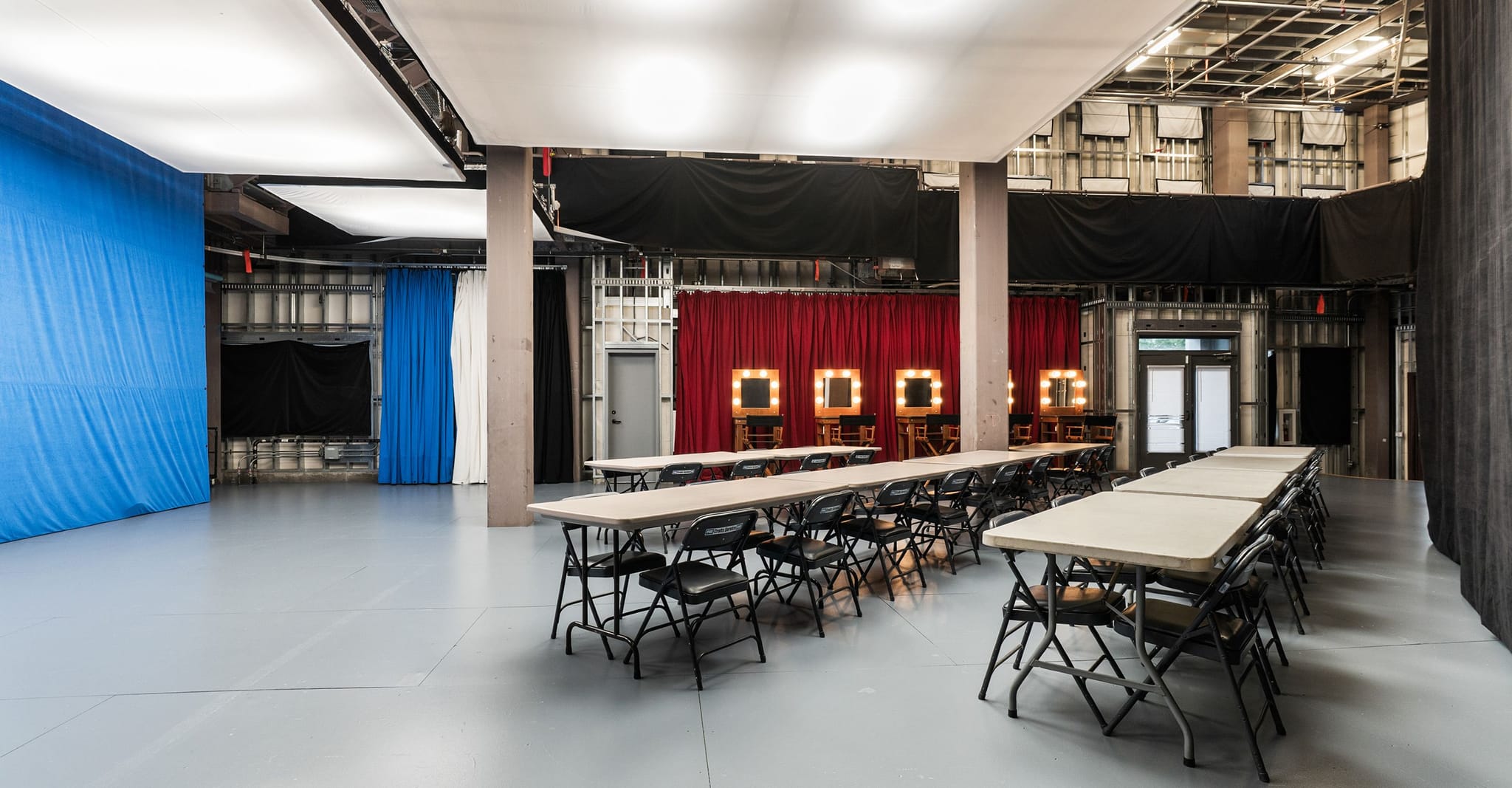 An expansive studio with a blue screen, rows of tables and chairs, and makeup stations under red drapes.