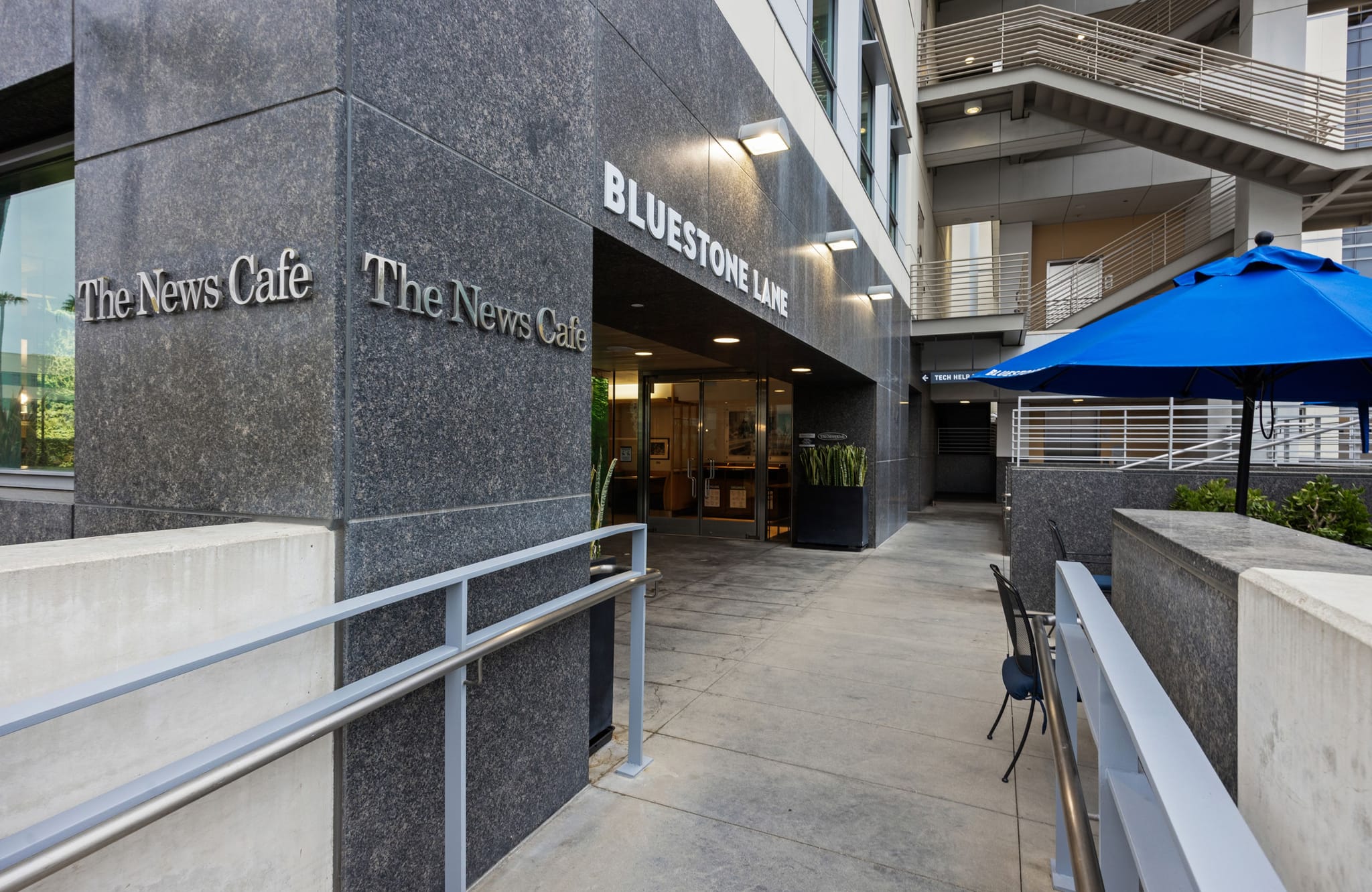 A dark granite building with "The News Cafe" and "Bluestone Lane" signs, a walkway leading to an entrance, and a blue patio umbrella with outdoor seating.