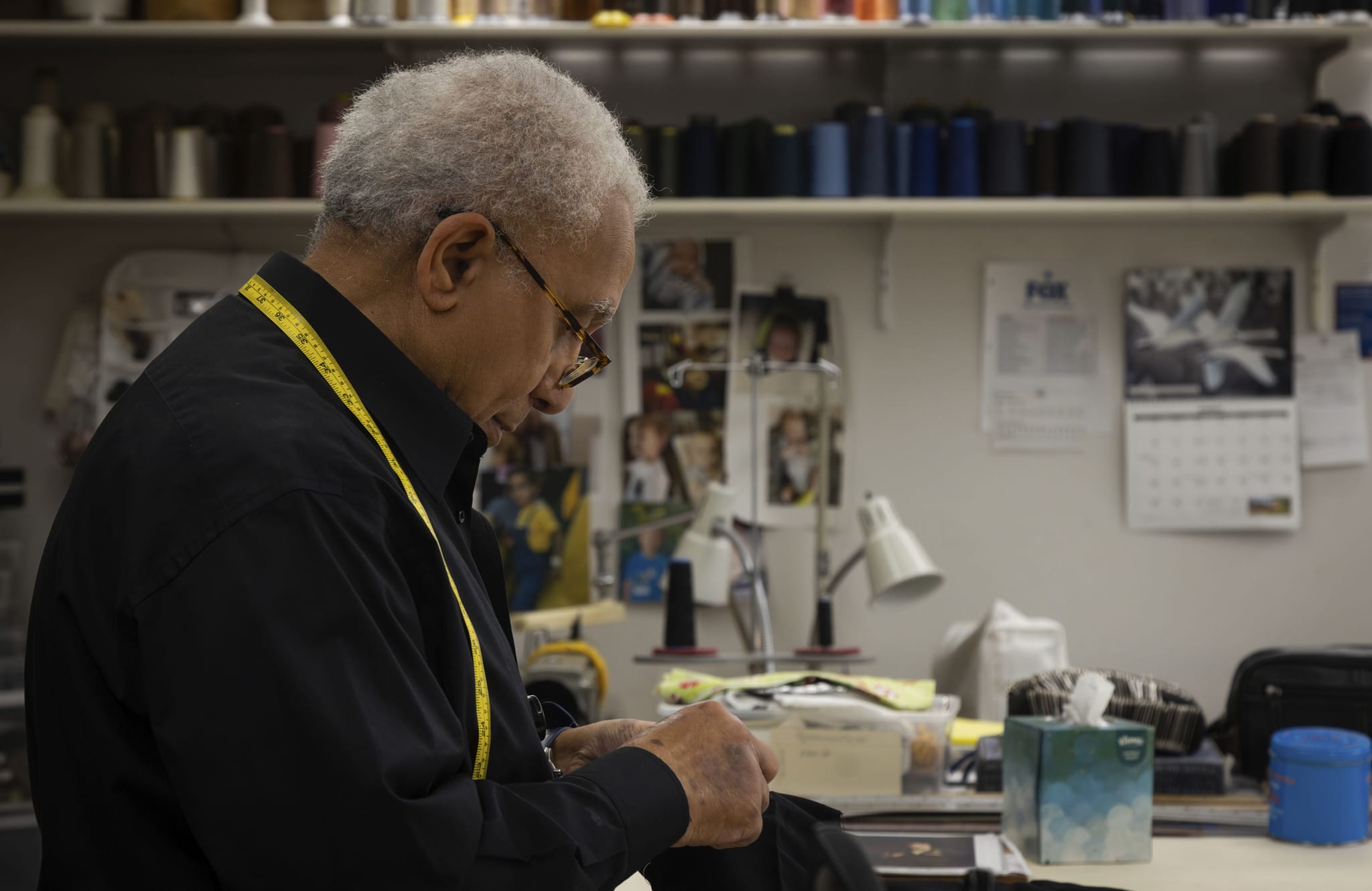 An older Black man with a measuring tape around his neck focuses on tailoring in a workshop.