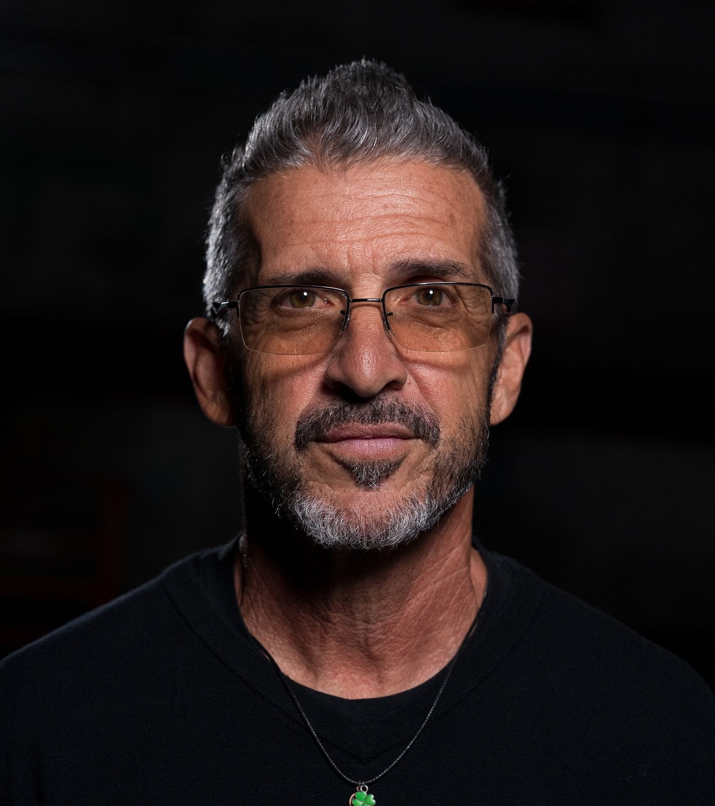 A man with spiked gray hair, a gray beard, and glasses wears a black shirt with a green pendant.