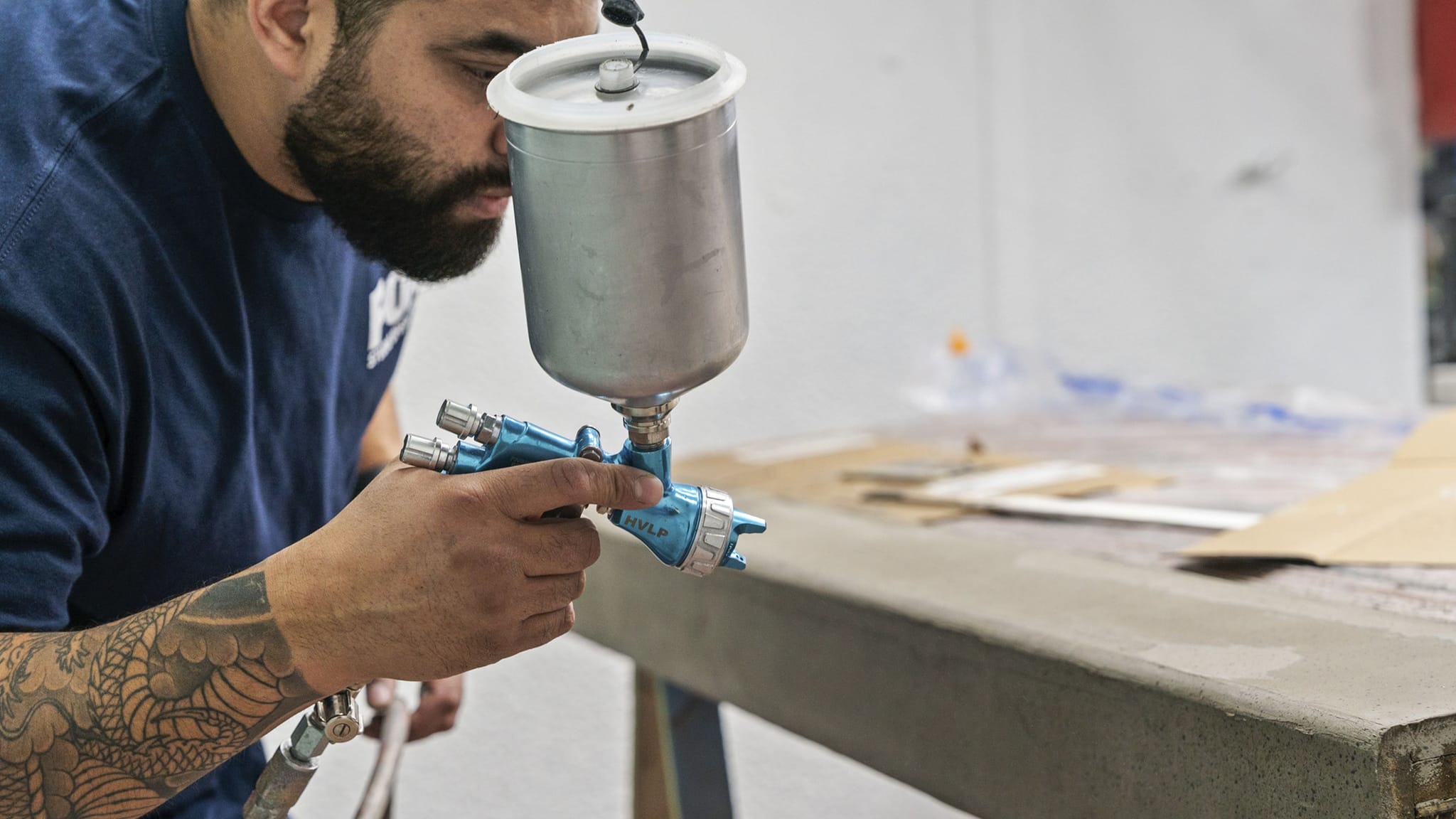 A bearded man with a tattooed arm holding a spray gun over a grey surface.