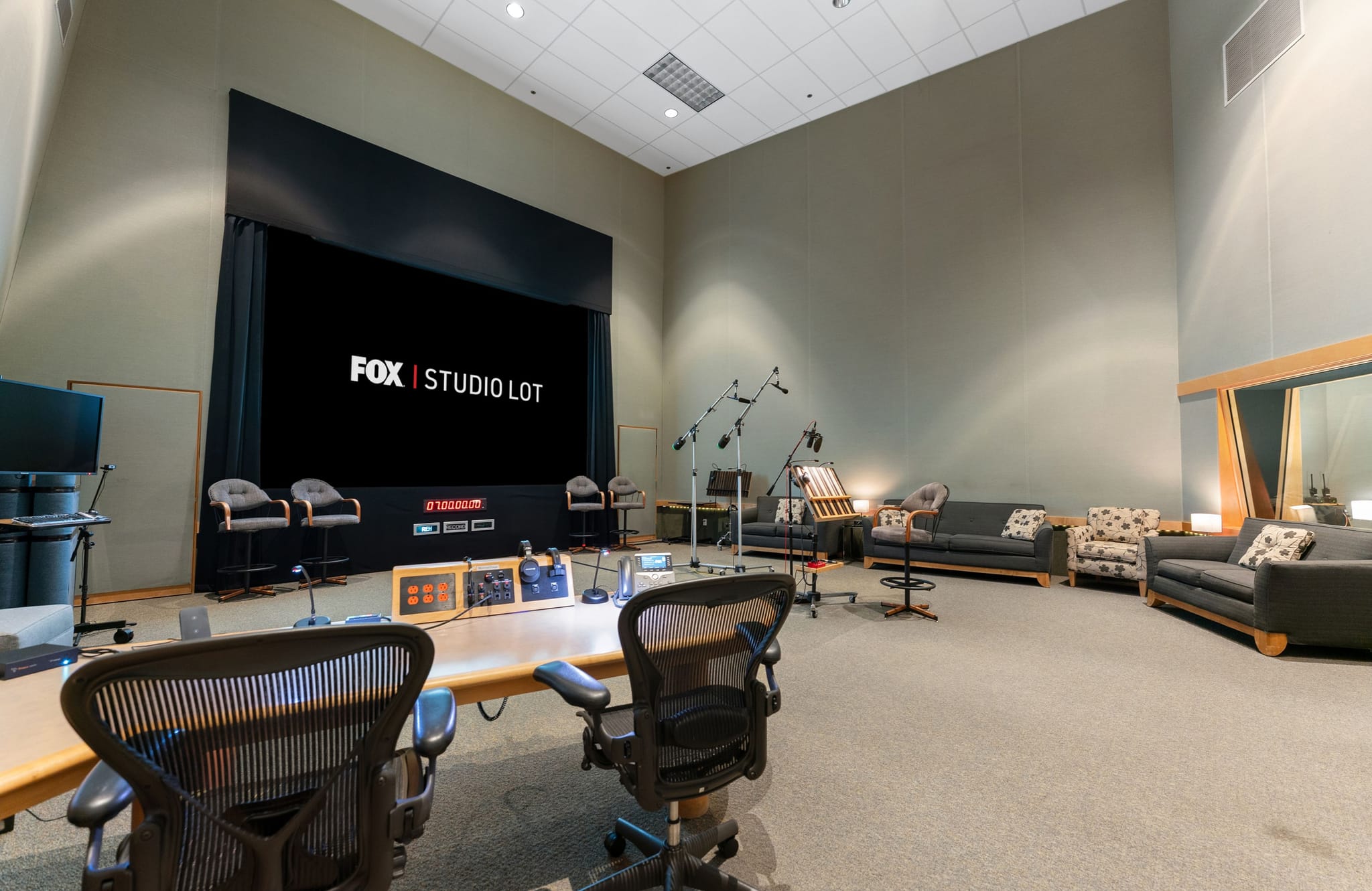 A recording studio with a large screen displaying "FOX | STUDIO LOT", a control desk, microphones, and lounge seating.