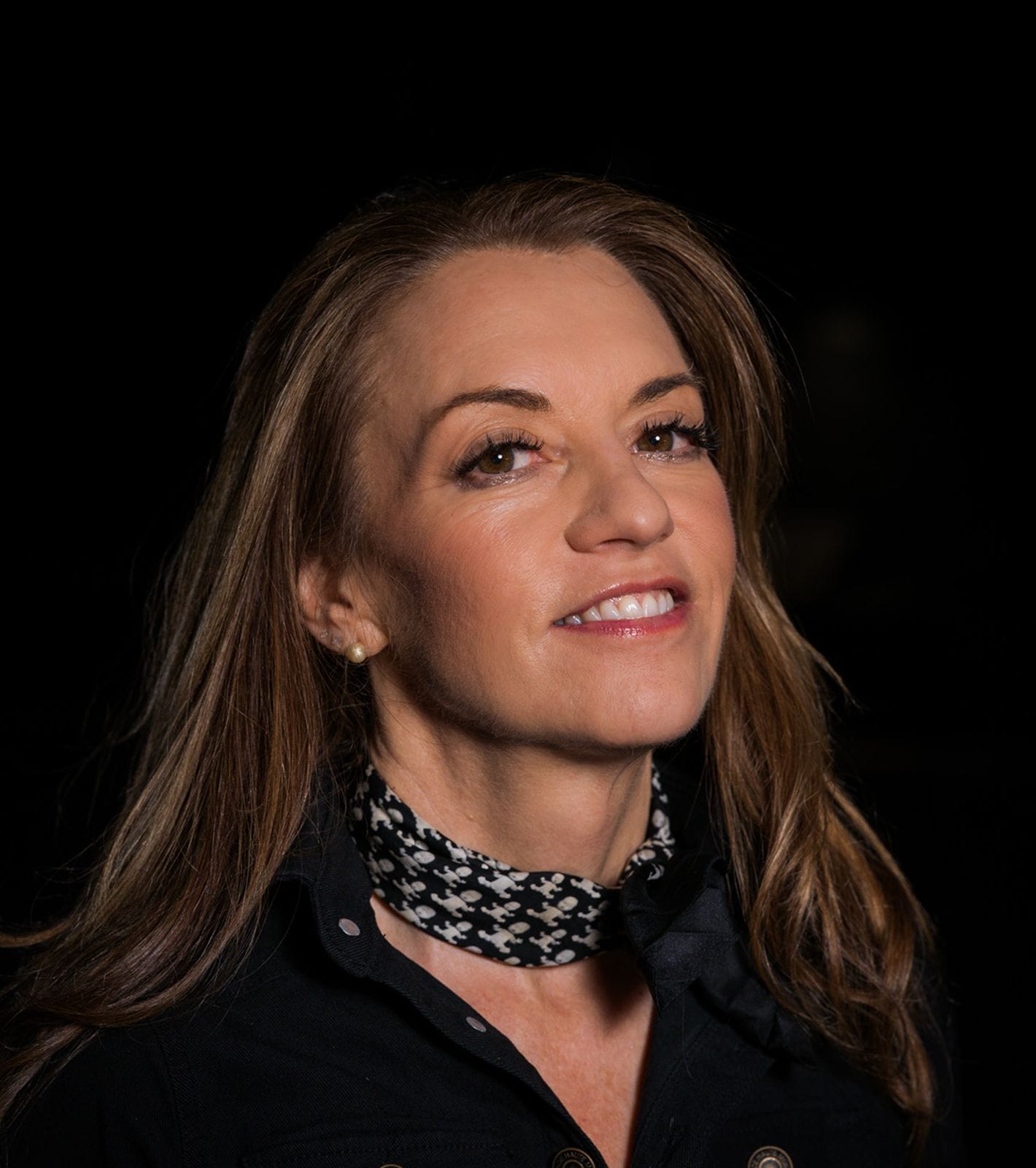 A woman with long brown hair, wearing a black shirt and a patterned bandana, smiles.