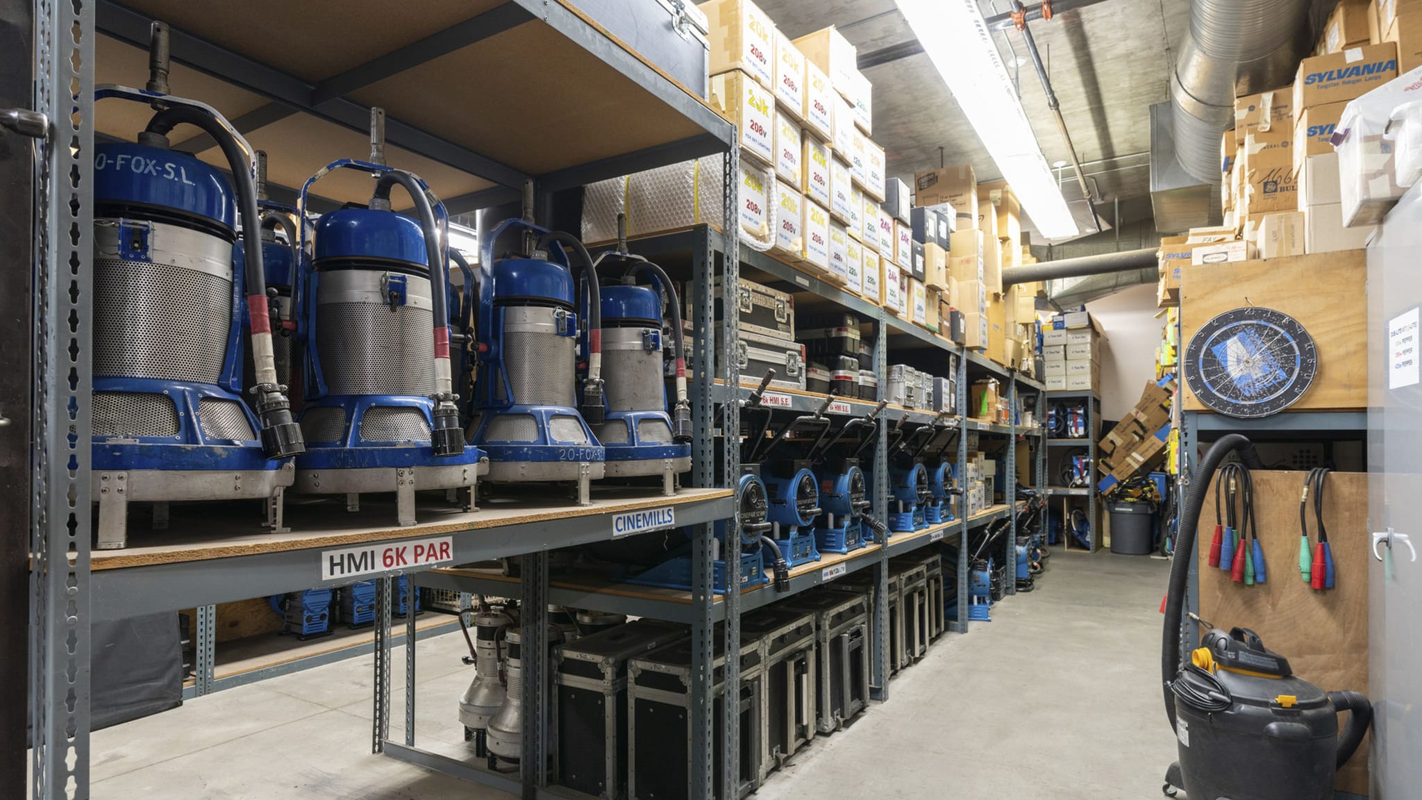 A storage room with metal shelves packed with film production equipment, including large blue HMI lights and numerous boxes.