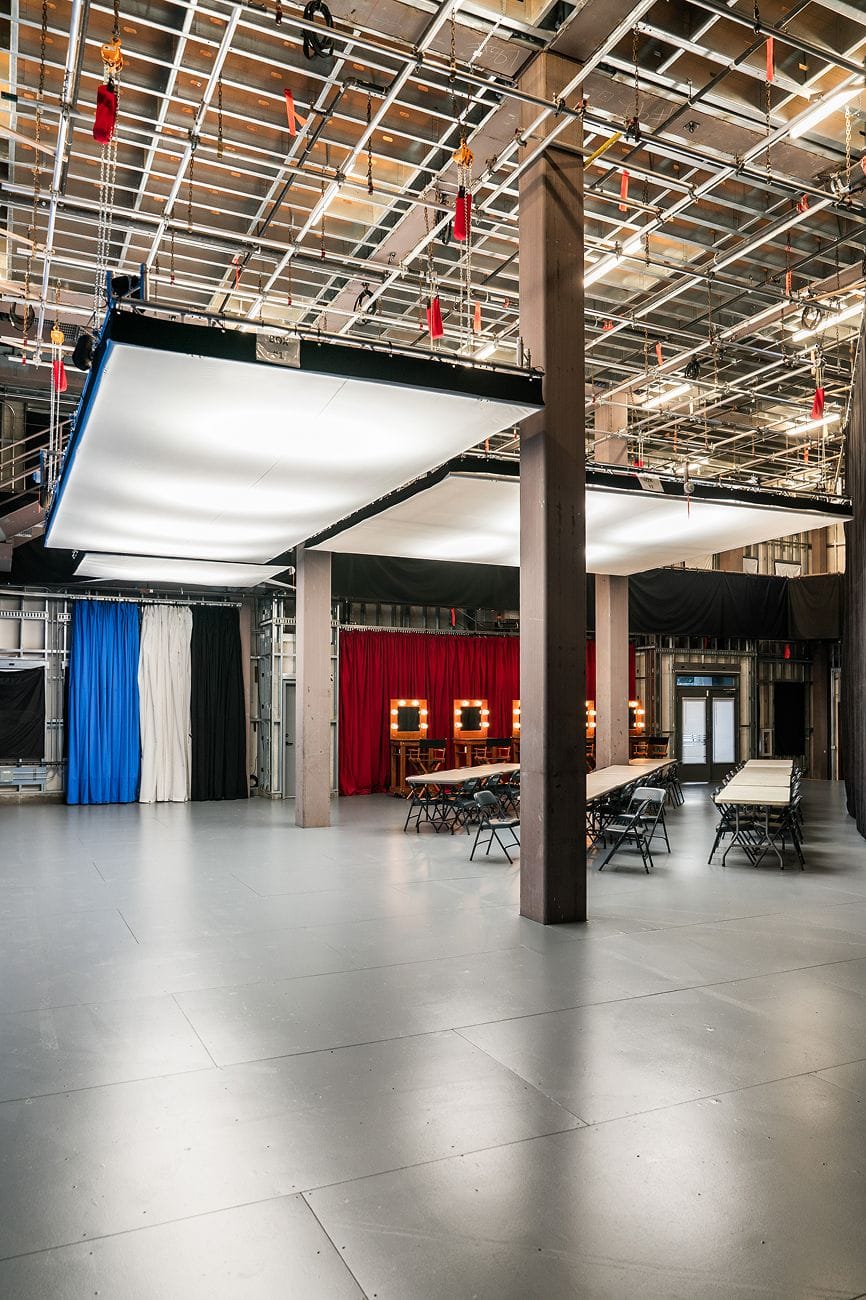 Spacious production studio with a grid ceiling, large overhead softboxes, colorful curtains, vanity mirrors, and long tables with chairs.