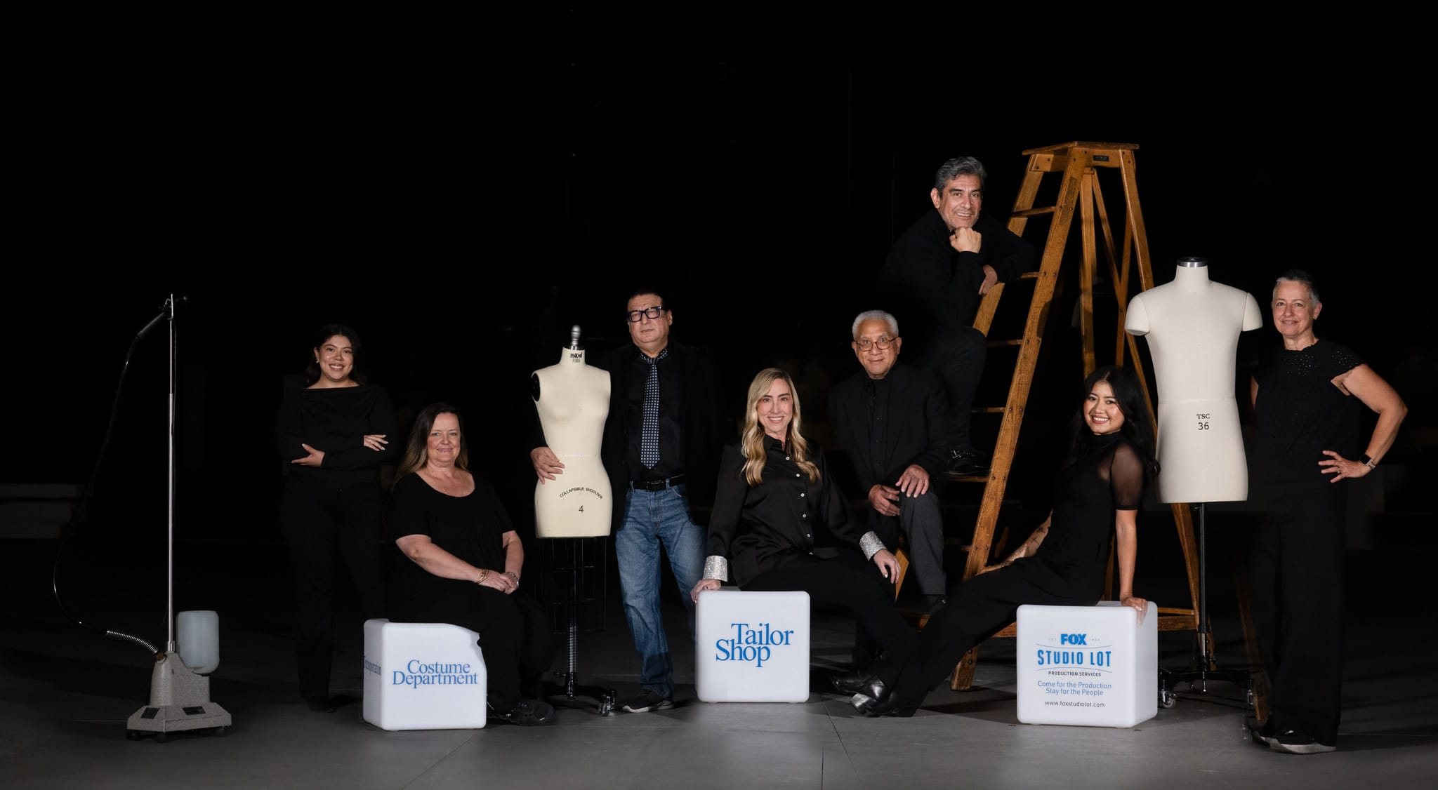 A team of costume and tailoring professionals poses in a studio with mannequins, a steamer, and cubes labeled 'Costume Department', 'The Tailor Shop', and 'FOX Studio 101'.
