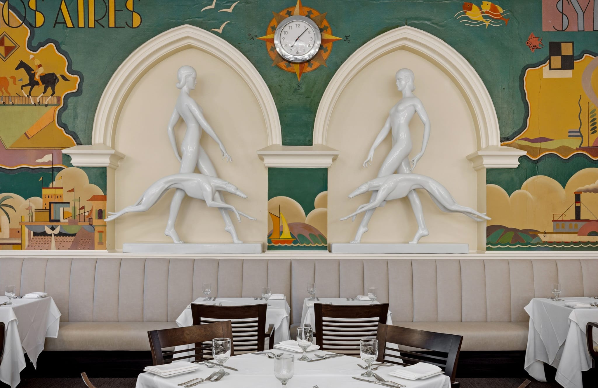 Art Deco dining room with a colorful mural, two white sculptures of figures with greyhounds, and a clock.