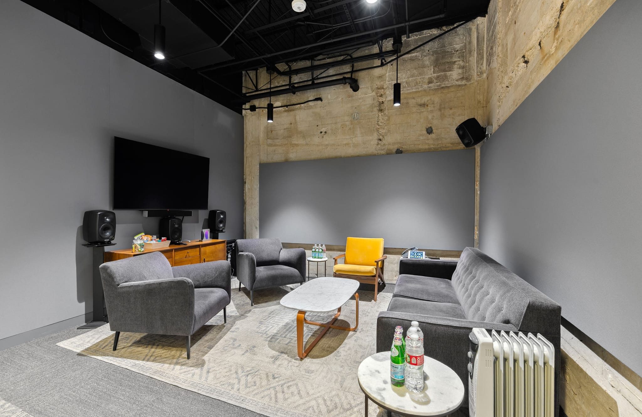 A modern industrial lounge with gray walls, a large TV, gray and yellow seating, and an exposed concrete ceiling.