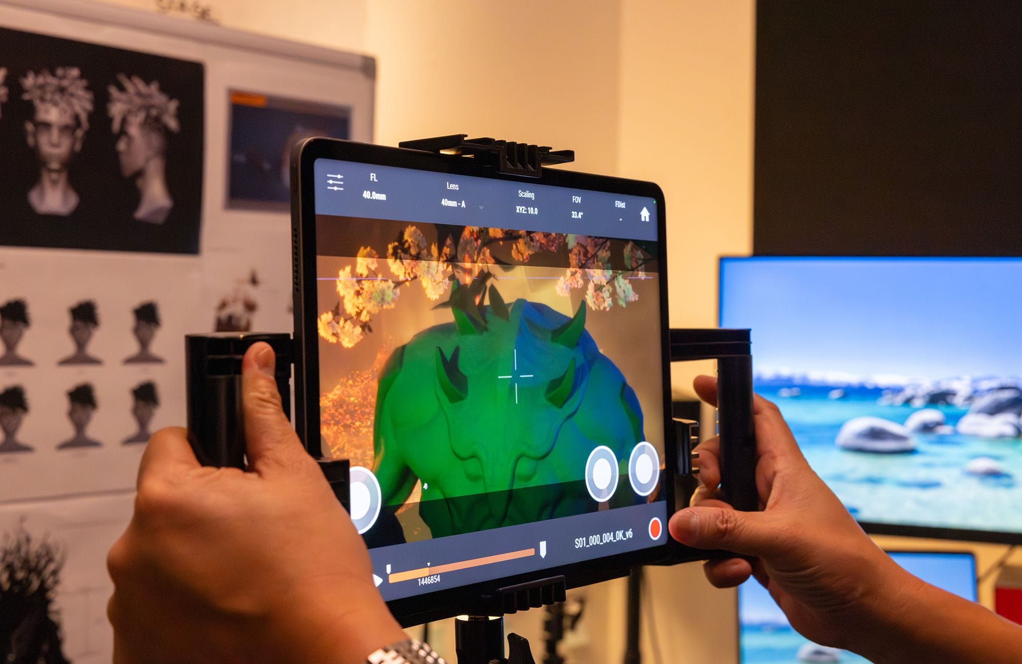 A person holds a tablet displaying a green and blue 3D creature model against a vibrant orange background, with studio concept art visible behind them.