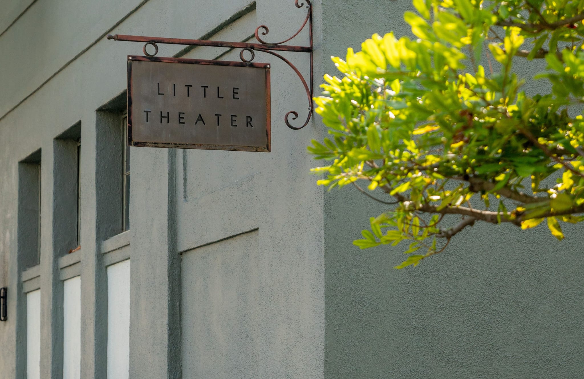 A dark metal "LITTLE THEATER" sign hangs on a light building, with green leaves on the right.