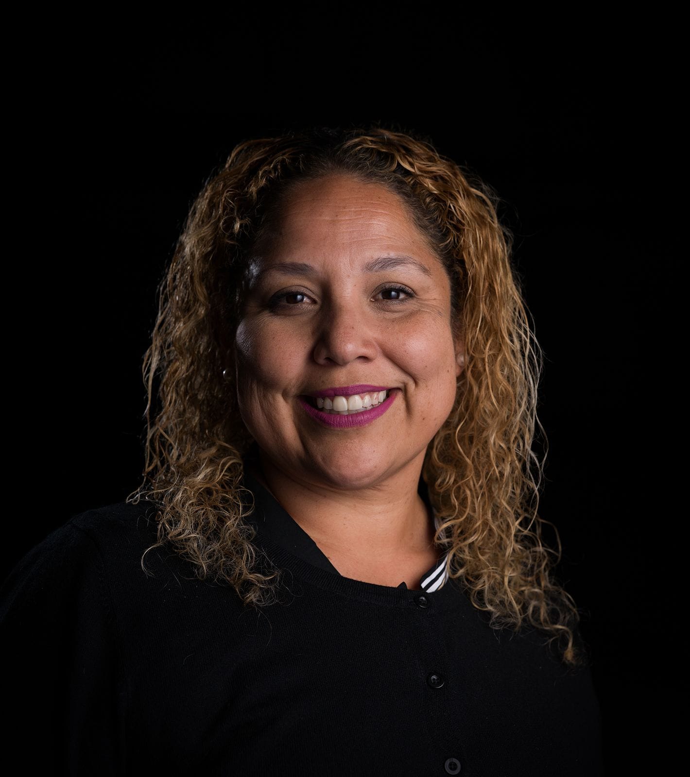 Smiling woman with curly light brown hair and pink lipstick against a black background.