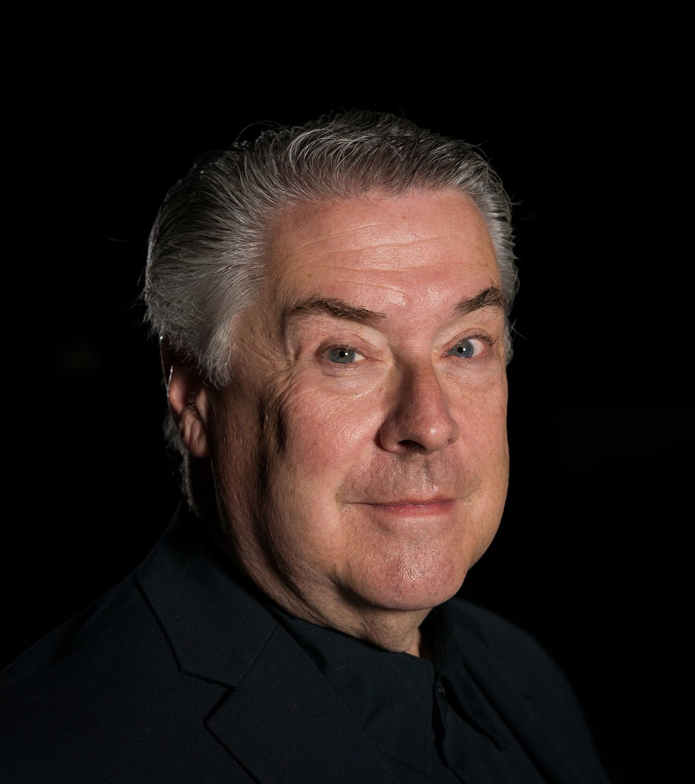 Headshot of an older man with grey hair smiling against a black background.