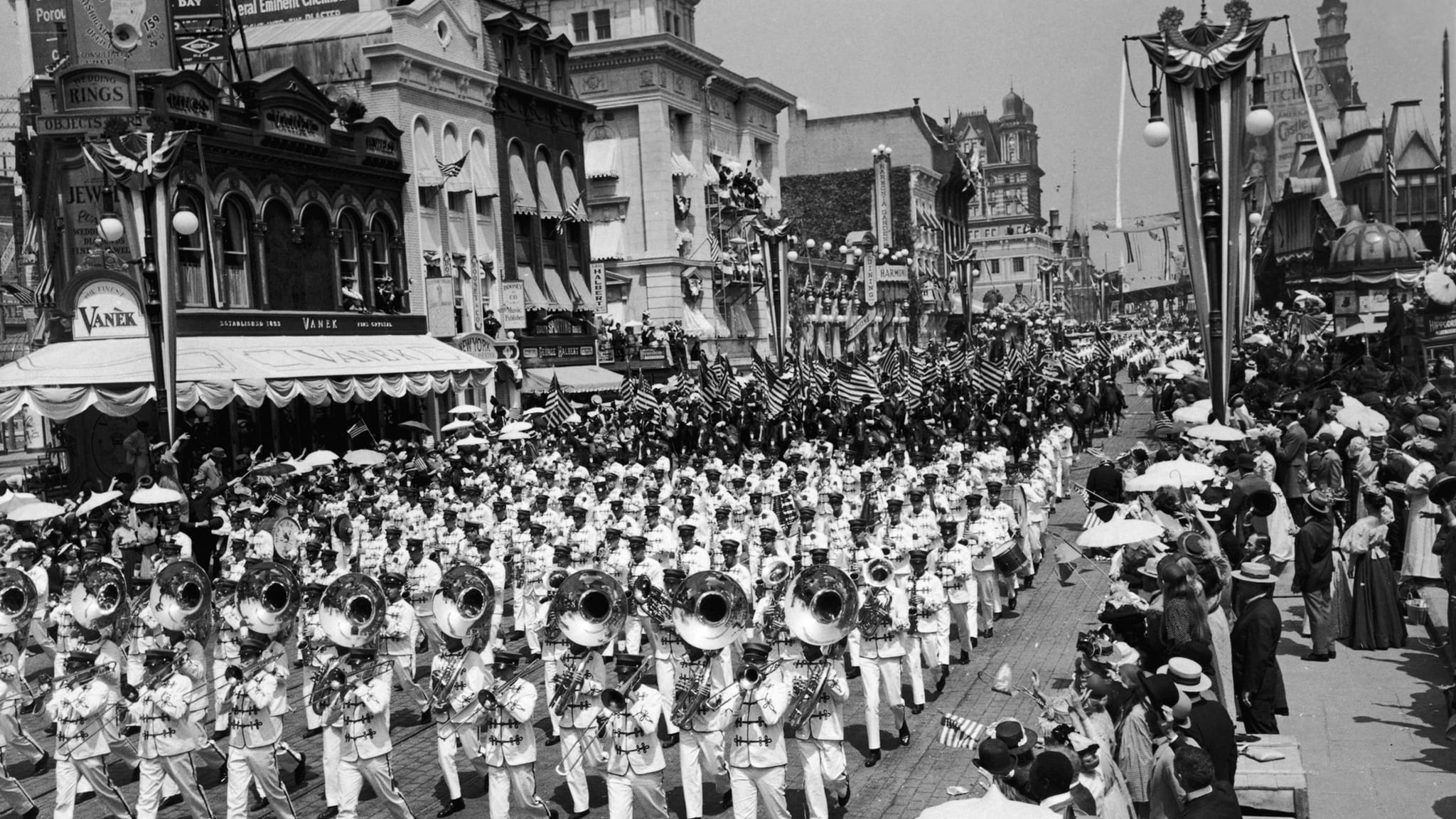Historic black and white photo of a large marching band leading a parade down a busy city street lined with spectators and decorated buildings.