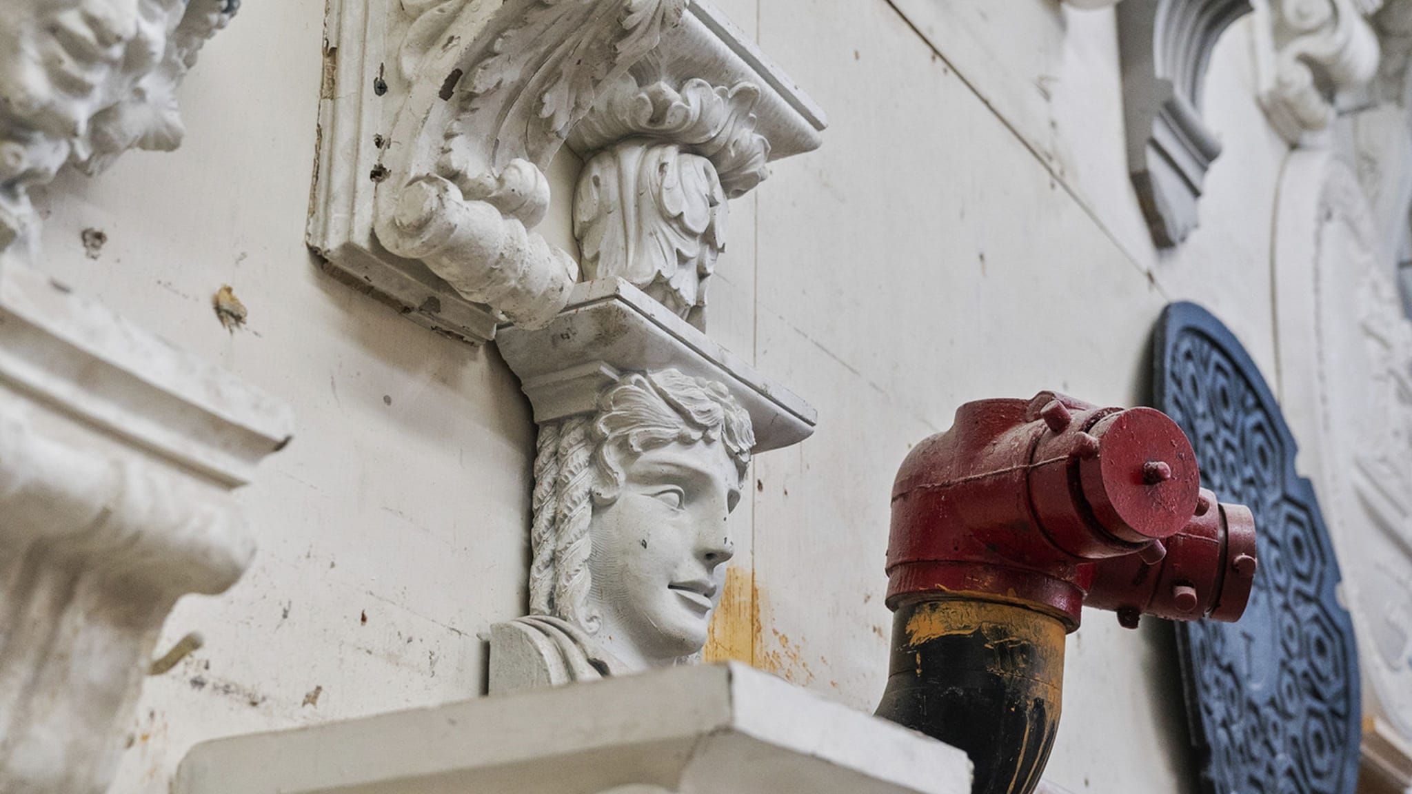 White classical architectural reliefs, including a bust and corbels, next to a red fire standpipe on a wall.
