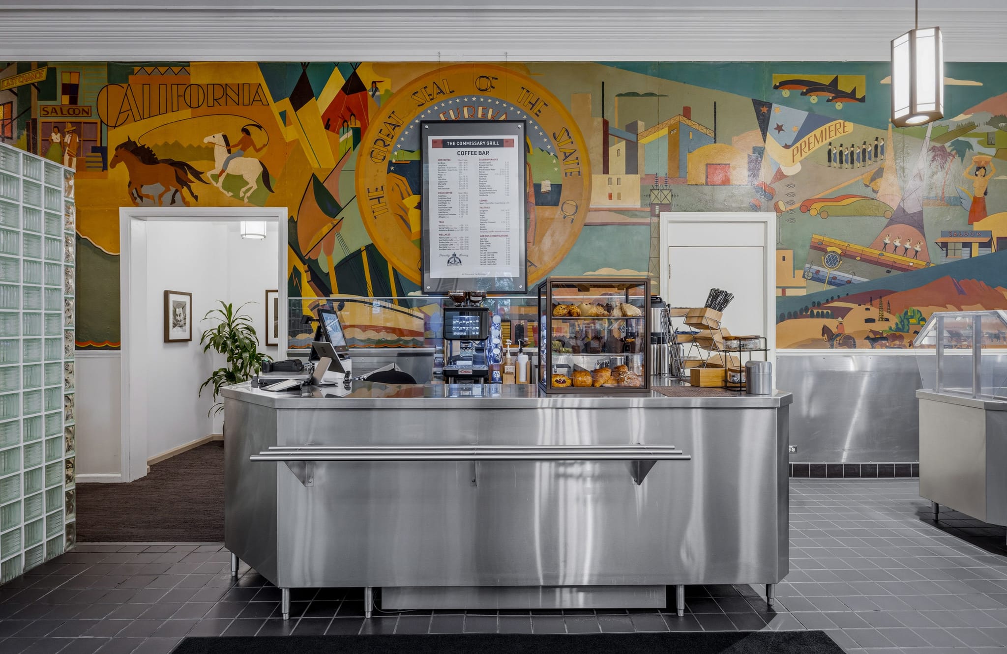 A cafe interior featuring a stainless steel coffee bar counter in front of a large, colorful mural depicting scenes of California history.
