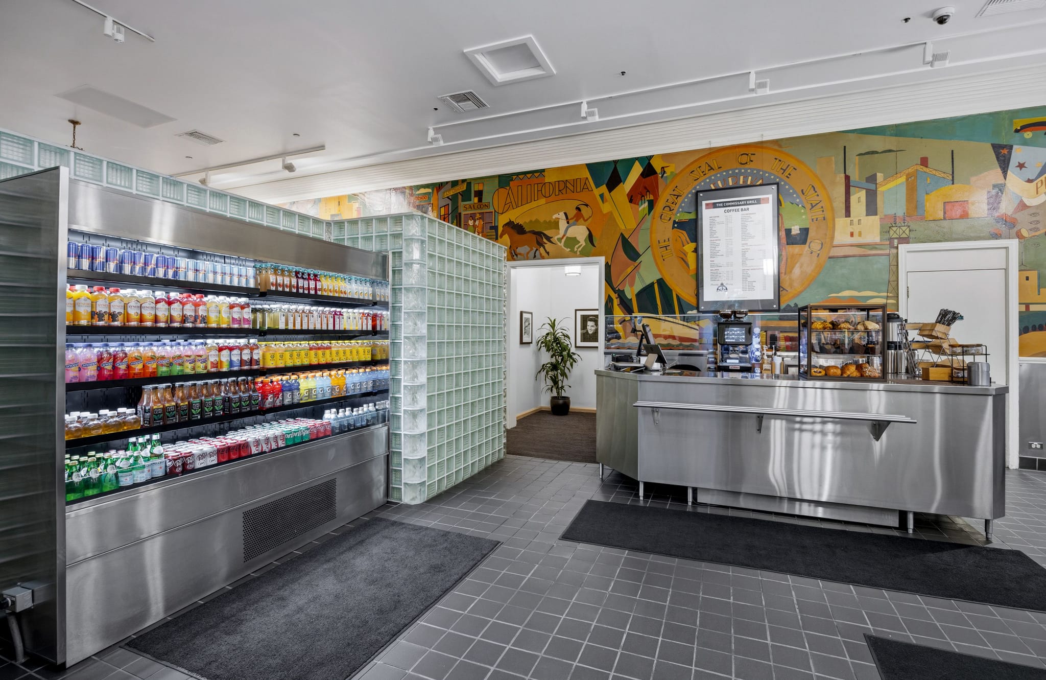 Interior of a modern deli with a refrigerated drink case, stainless steel service counter, and a colorful mural on the back wall.