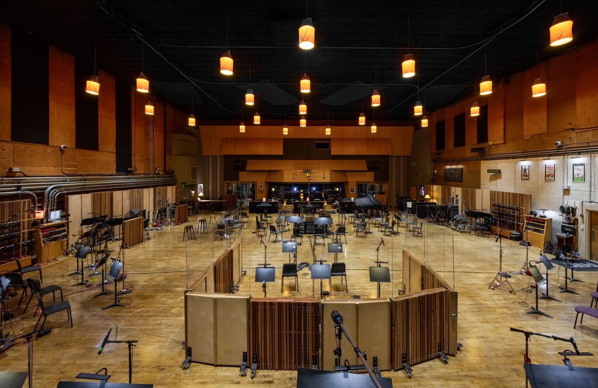 An empty, spacious recording studio with wooden floors, many chairs, music stands, sound baffles, and warm pendant lights.