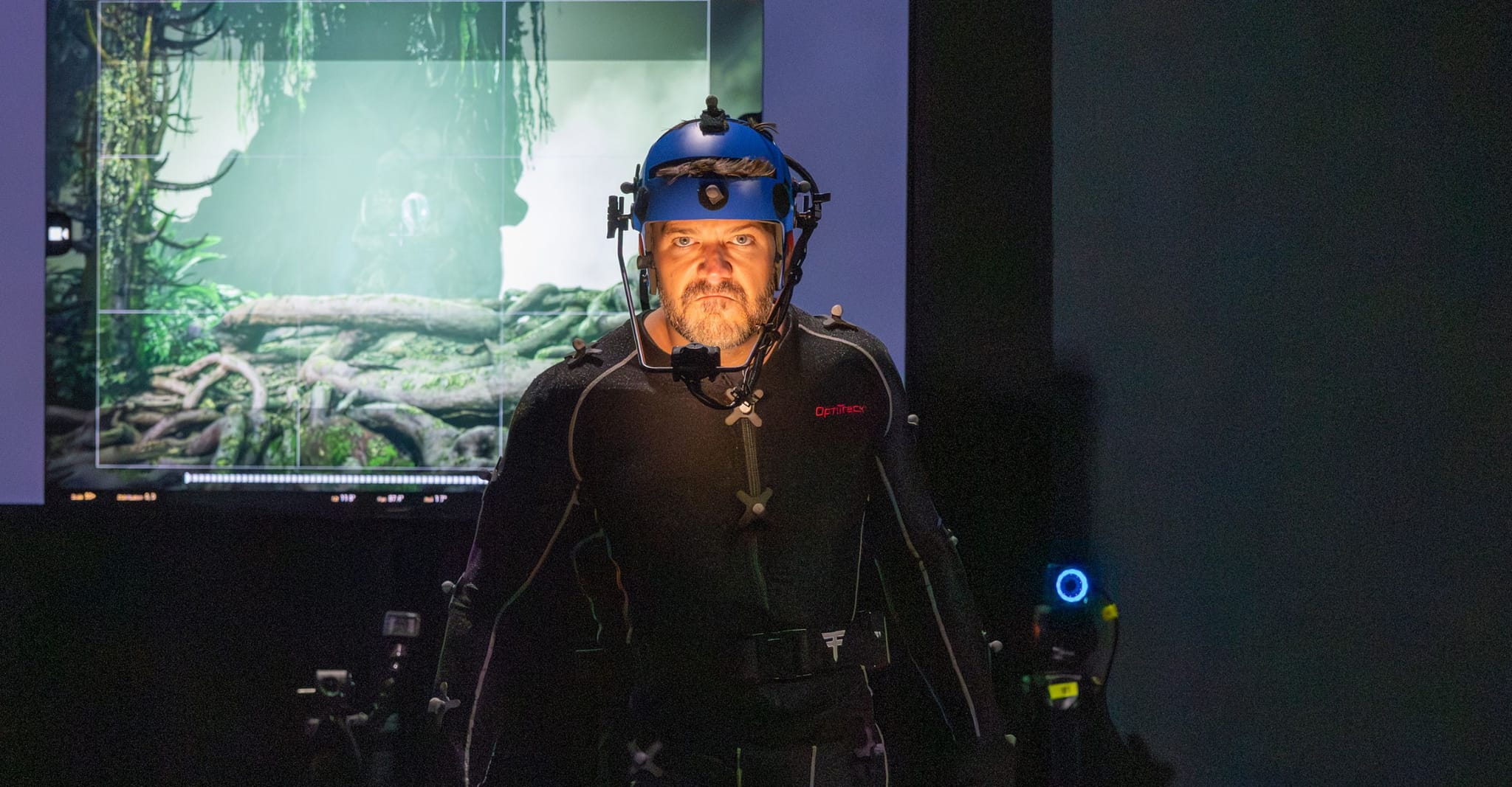 A man in a motion capture suit and helmet stands in front of a screen displaying a jungle scene.
