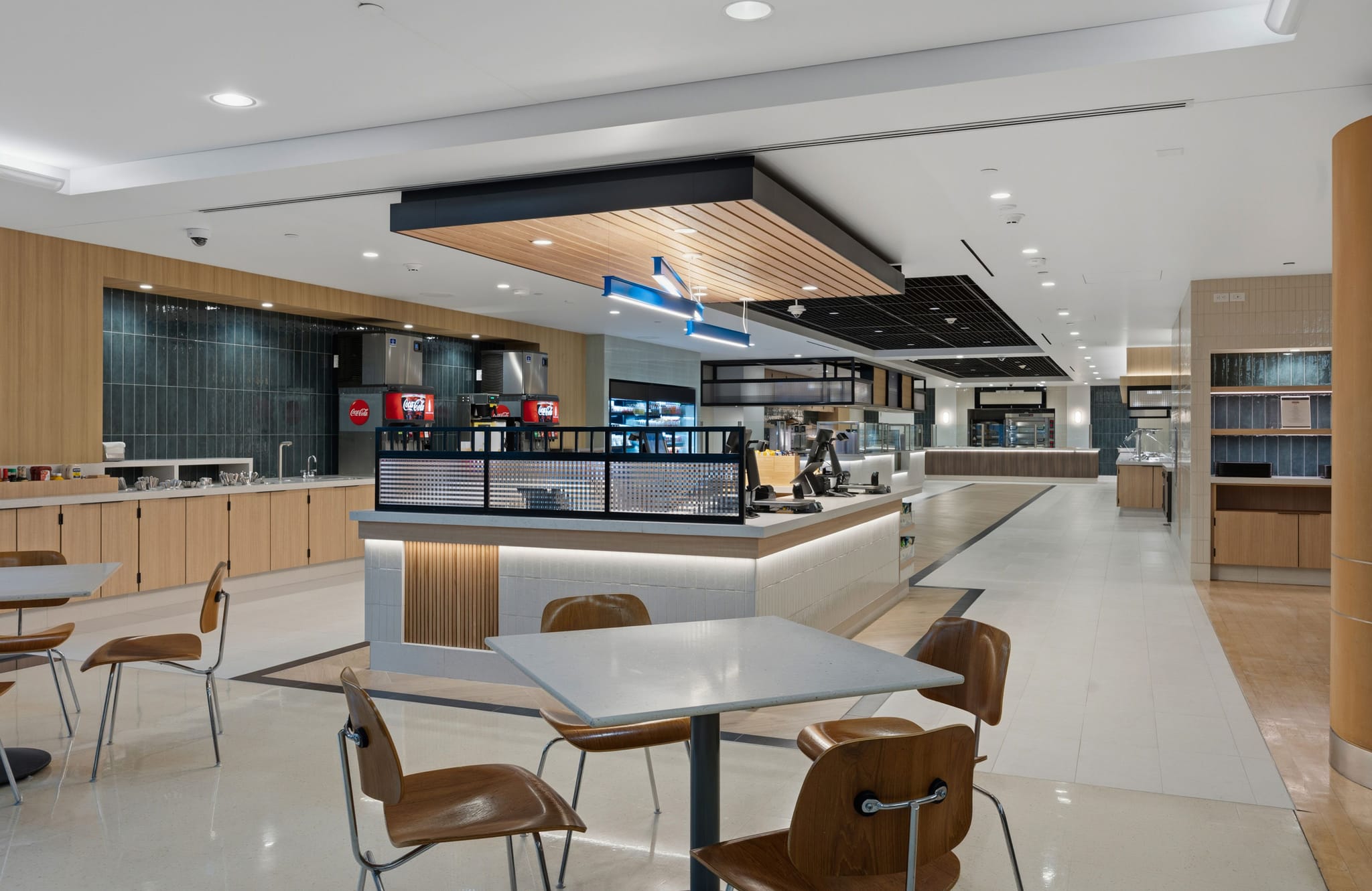 A spacious, modern cafeteria with light wood walls, dark tile accents, a central food counter, and scattered white tables with brown chairs.