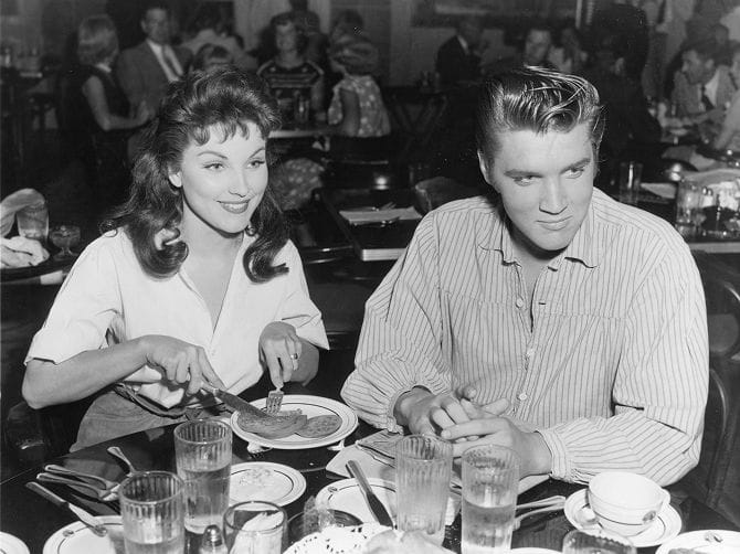 Black and white photo of Elvis Presley and Ann-Margret seated at a diner table. She smiles while cutting food, and Elvis looks to the side with clasped hands.