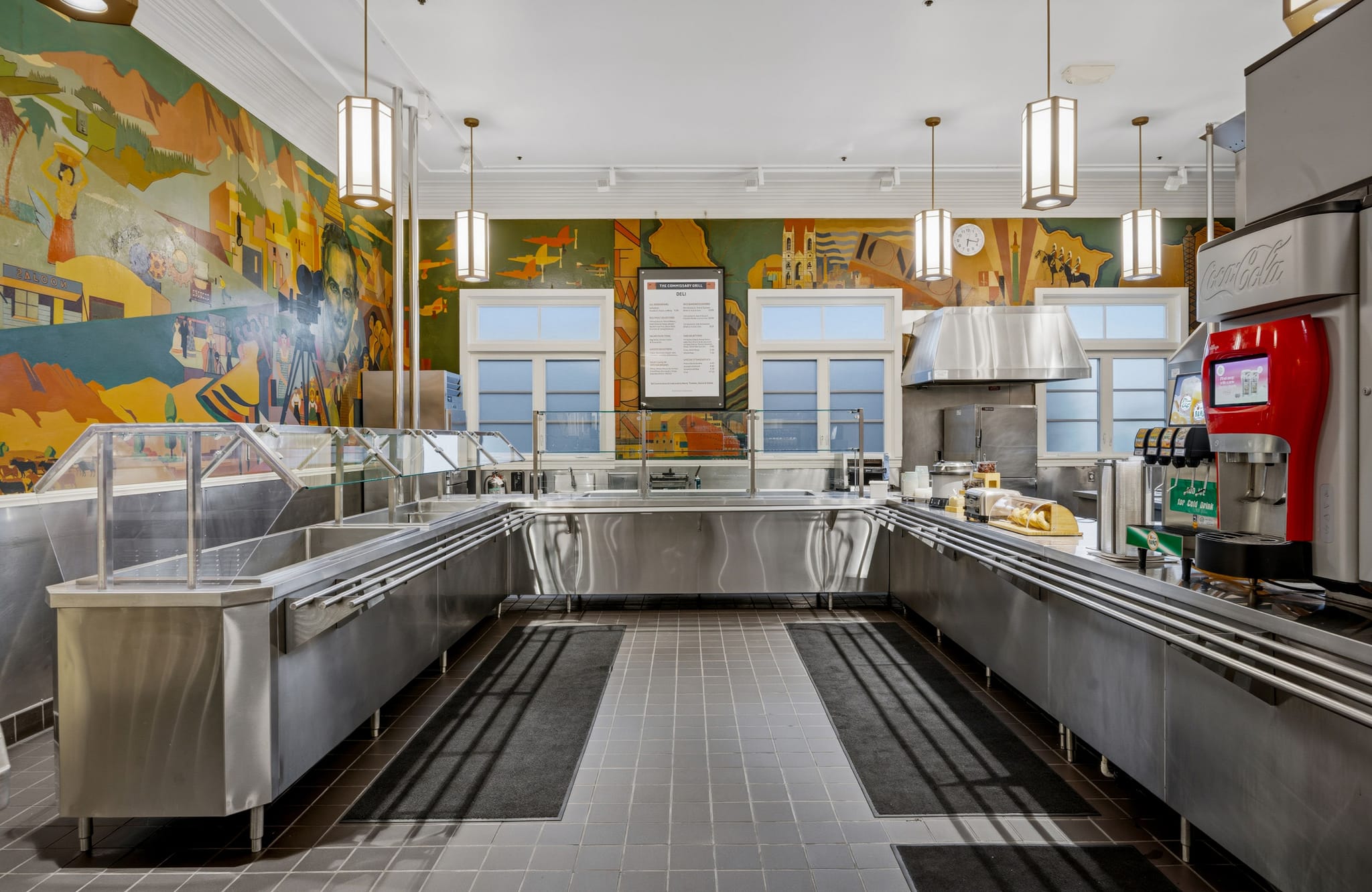 An empty cafeteria serving line with U-shaped stainless steel counters and a vibrant mural on the back wall.